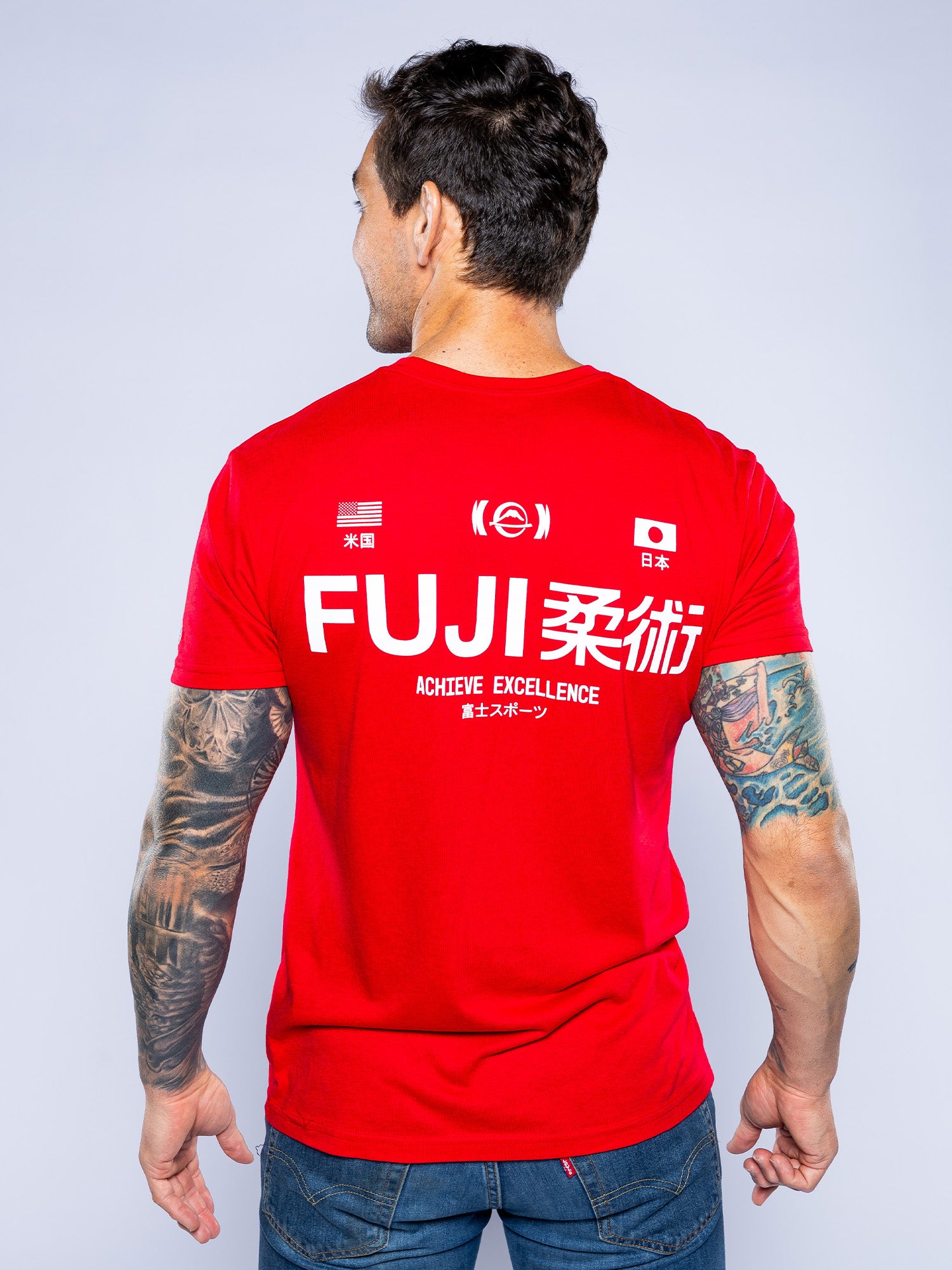 A tattooed man faces away, wearing the Iconic Jiu Jitsu Shirt Red with "FUJI," American and Japanese flags, and "ACHIEVE EXCELLENCE" in white text on the back.
