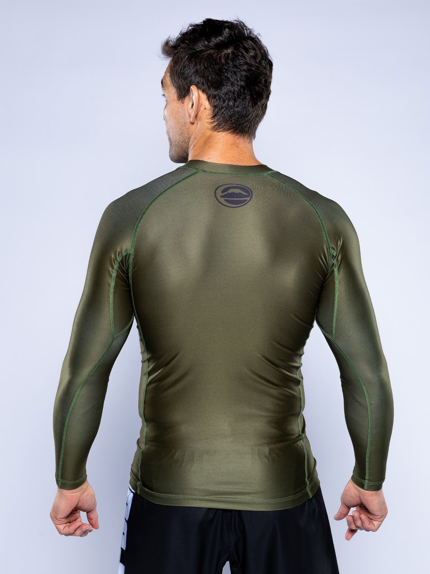 A man faces away, wearing the Baseline 2.0 Long Sleeve Rashguard in Military Green with dark green stitching, black shorts, and a small circular logo on the upper back. The background is plain and light-colored.