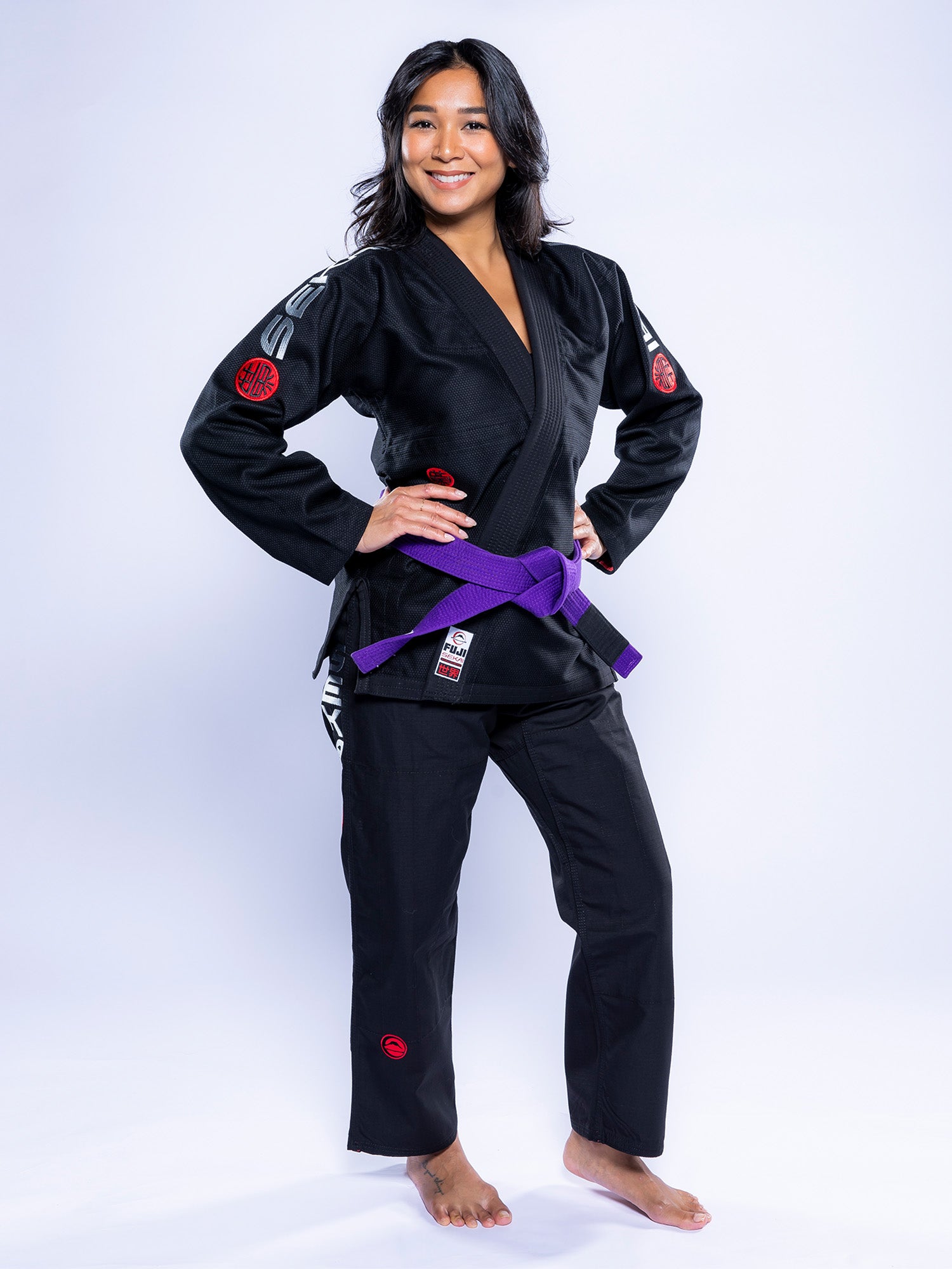 A woman stands barefoot on a white background, smiling with her hands on her hips and wearing the Womens SEKAI 3.0 BJJ Gi Black Grey with a purple belt.