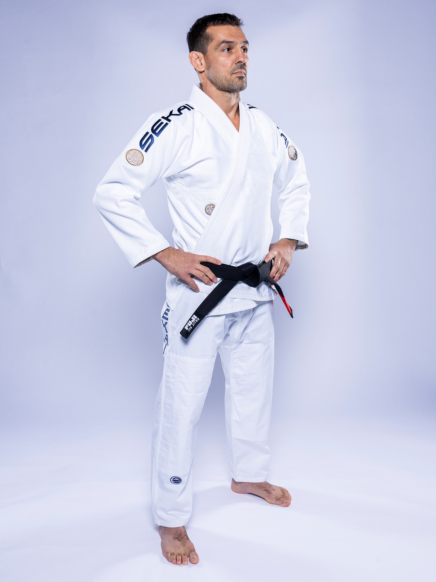 A man in a SEKAI 3.0 BJJ Gi White Navy and a black belt stands barefoot with hands on hips, gazing to the side against a light background.
