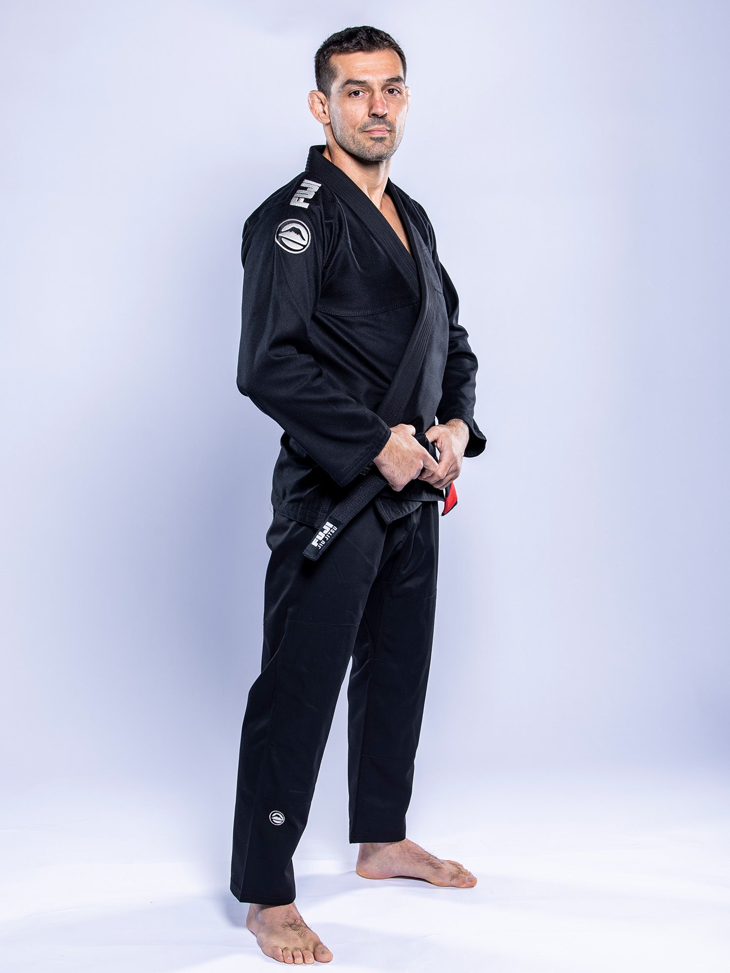 A man stands barefoot on a white background, confidently facing slightly to the side and holding his belt while wearing the Baseline 2.0 BJJ Gi Black Grey.