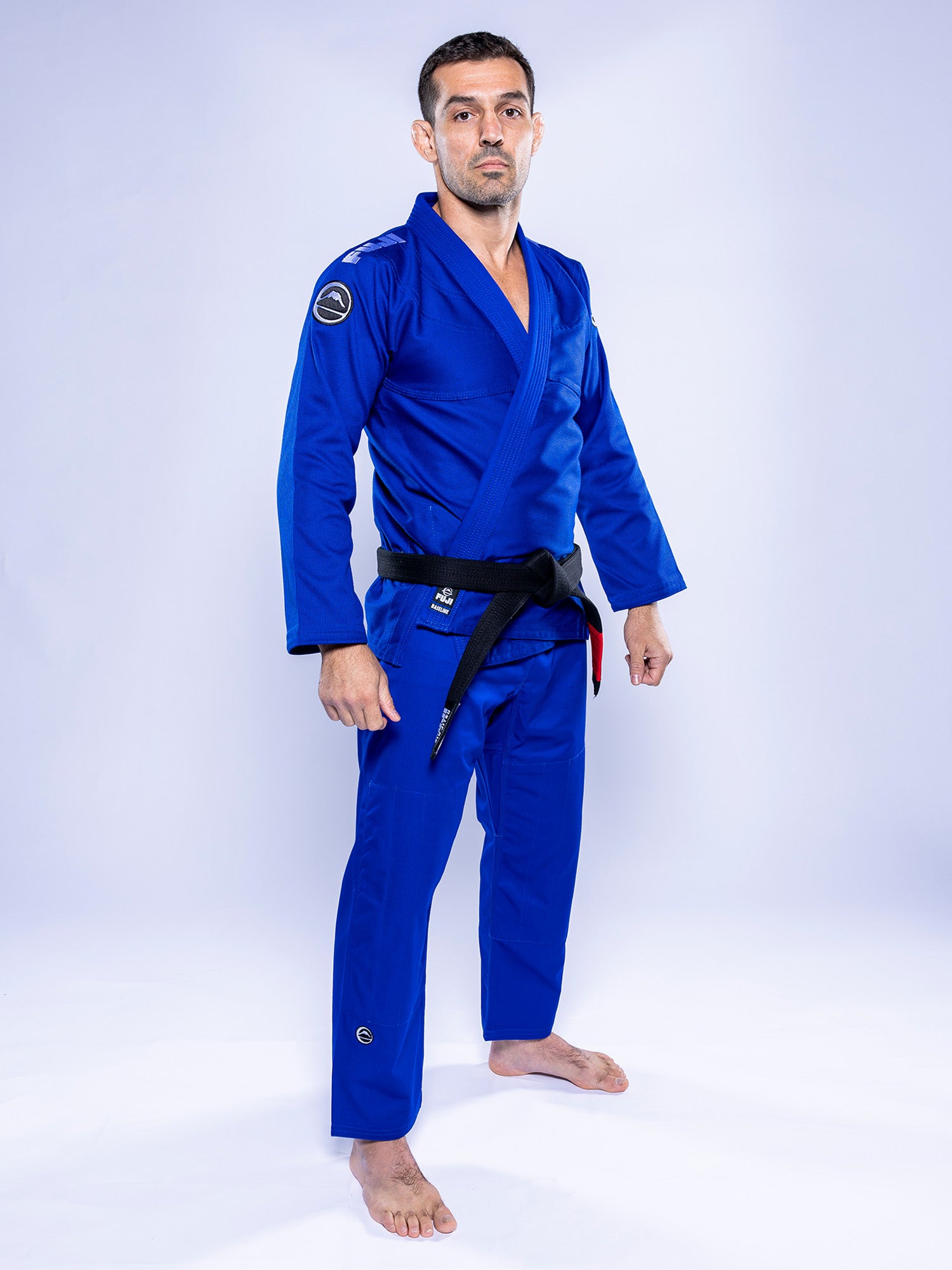 A man stands barefoot on a white background wearing the Baseline 2.0 BJJ Gi Blue White and a black belt, looking confidently at the camera.