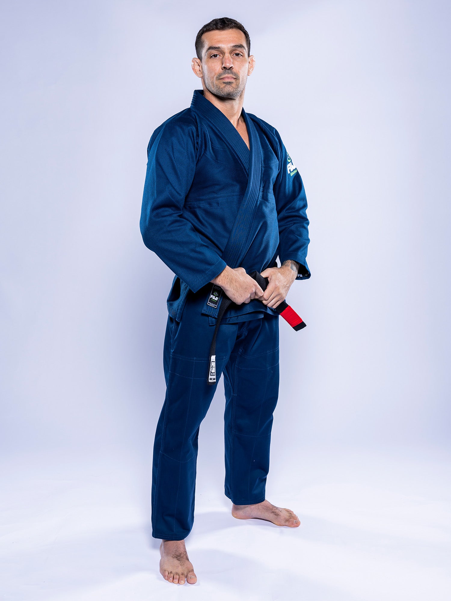 A man stands barefoot on a white background, wearing the Lightweight BJJ Gi Navy and holding a red belt in front of him, looking confidently at the camera.