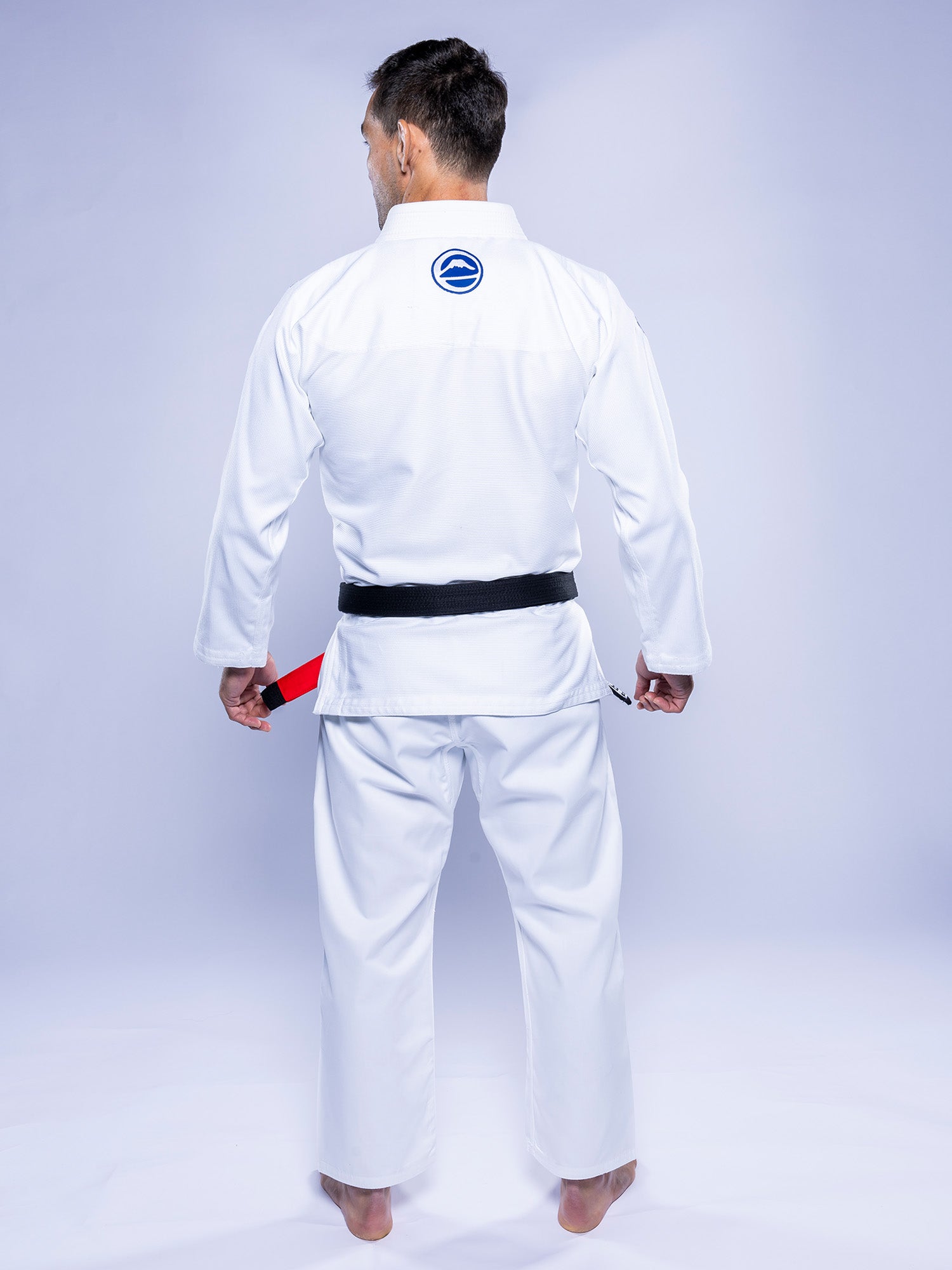 A person stands barefoot facing away, wearing the Baseline 2.0 BJJ Gi White Navy with a black belt. The uniform features a blue circular logo on the back and the individual's hands are at their sides.