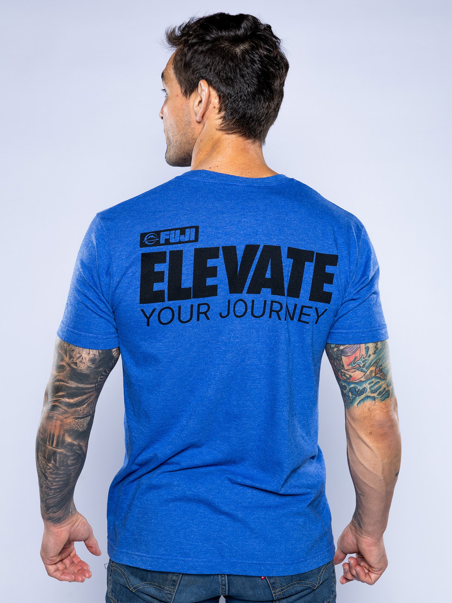 A man with tattooed arms faces away, wearing the Elevate Your Journey Shirt Blue featuring black "FUJI ELEVATE YOUR JOURNEY" text on the back. The background is plain and light-colored.