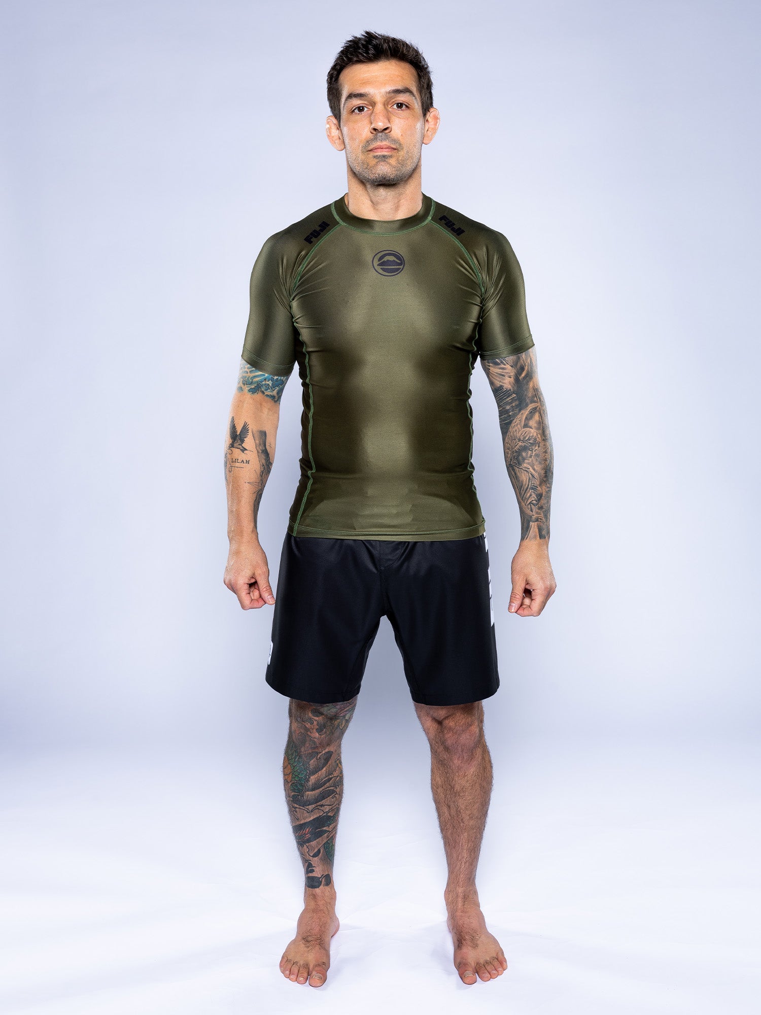 A man with tattoos stands barefoot on a white background, wearing the Baseline 2.0 Short Sleeve Rashguard in Military Green and black shorts, facing forward with arms relaxed at his sides.
