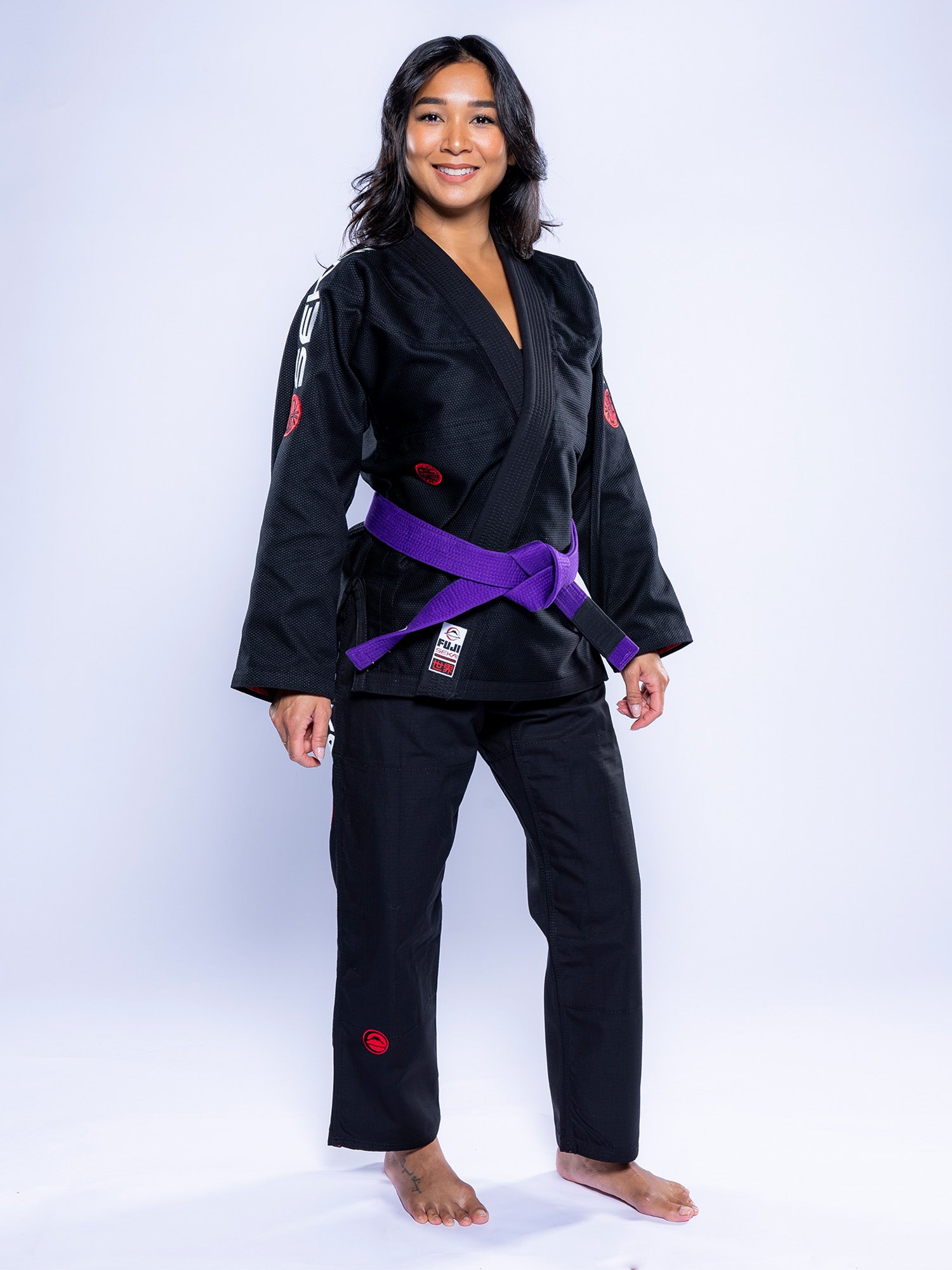 A woman stands barefoot in a Womens SEKAI 3.0 BJJ Gi Black Grey with a purple belt, smiling at the camera against a plain white background.