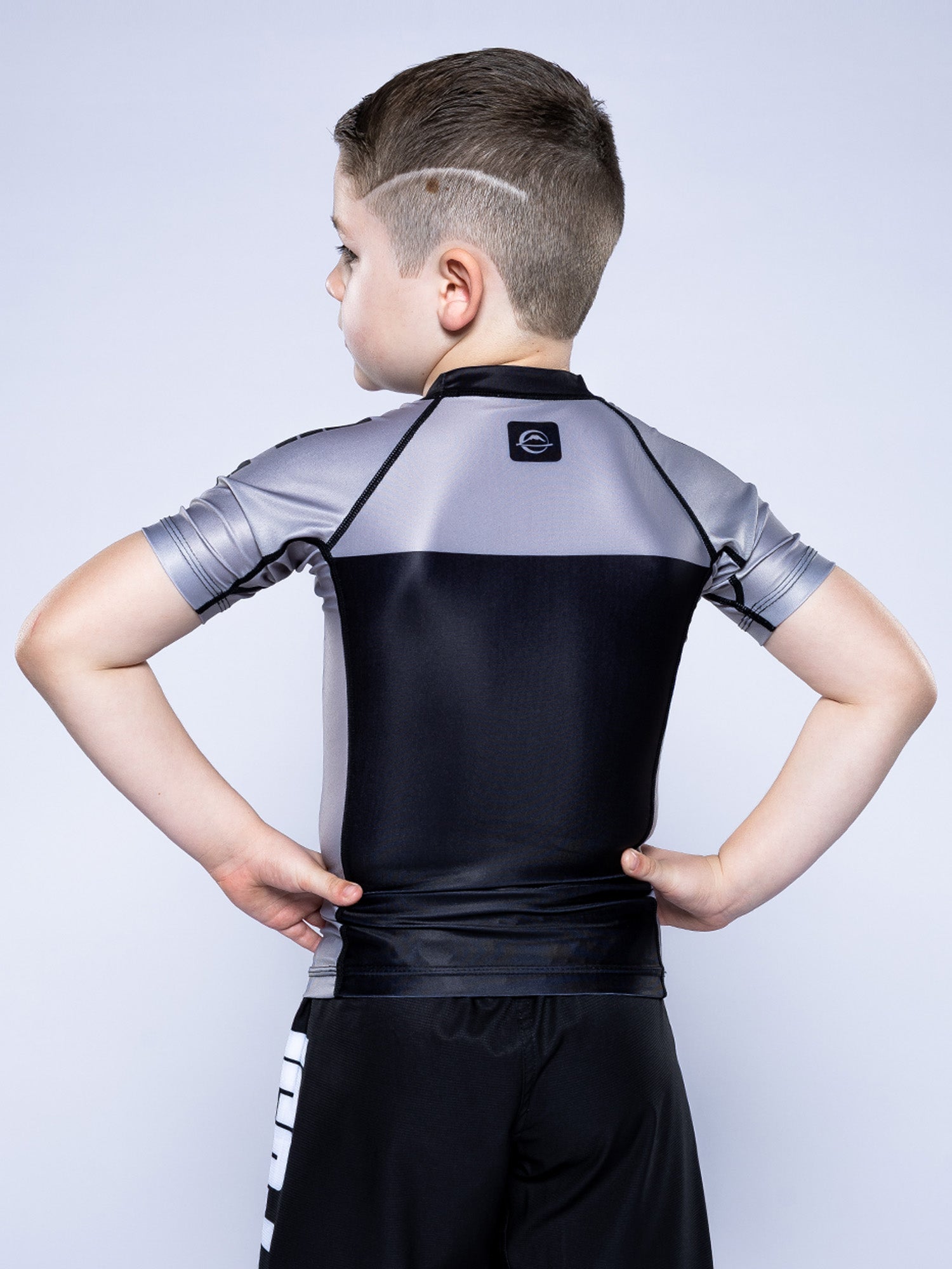 A young boy, seen from behind with hands on hips, wears the Kids Competition Ranked Rashguard Grey and black shorts. He has a modern haircut with trimmed sides, standing against a plain light background.