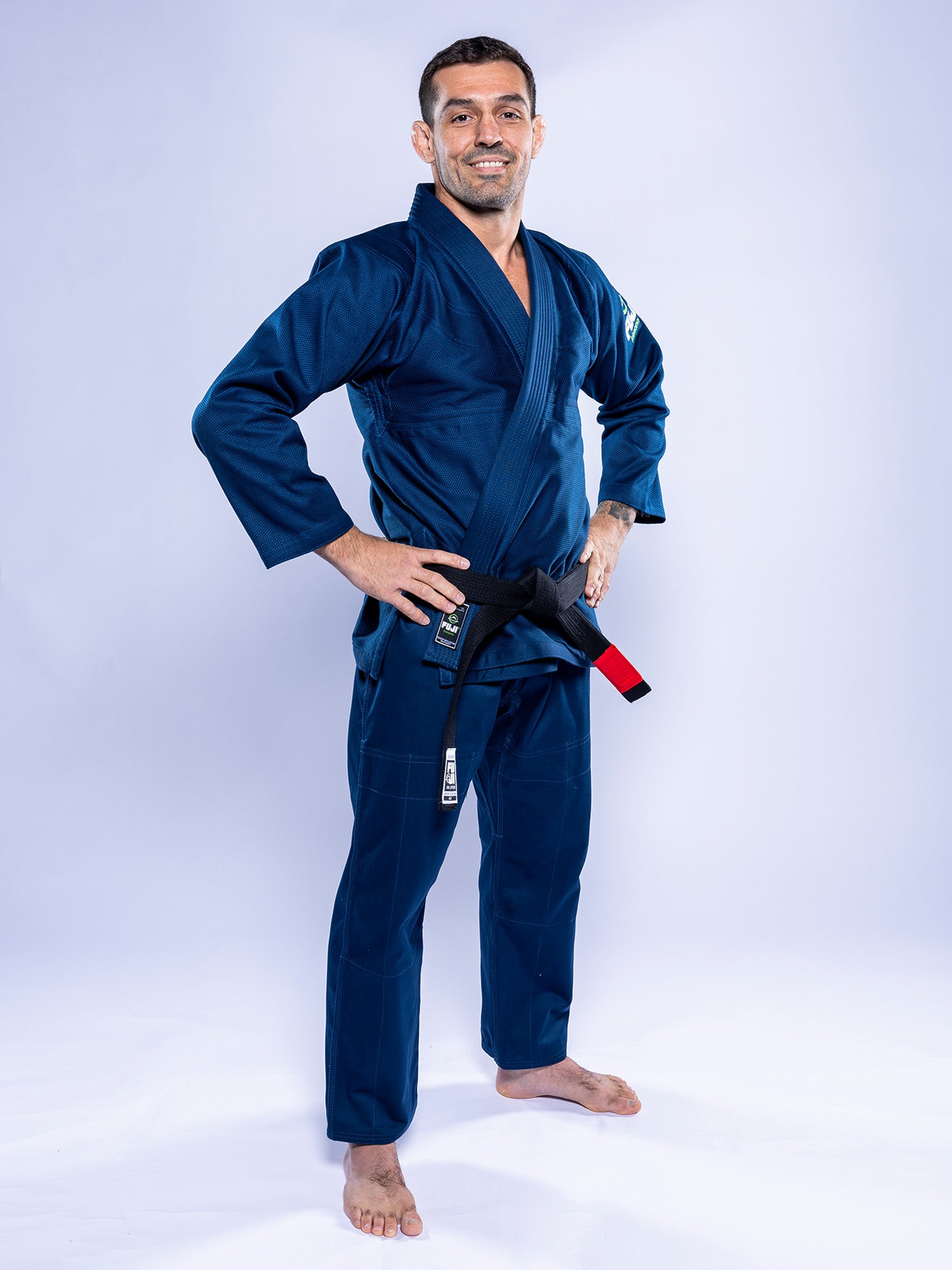 A man stands barefoot on a white background, smiling with his hands on his hips while wearing the Lightweight BJJ Gi Navy and a black belt with a red bar.