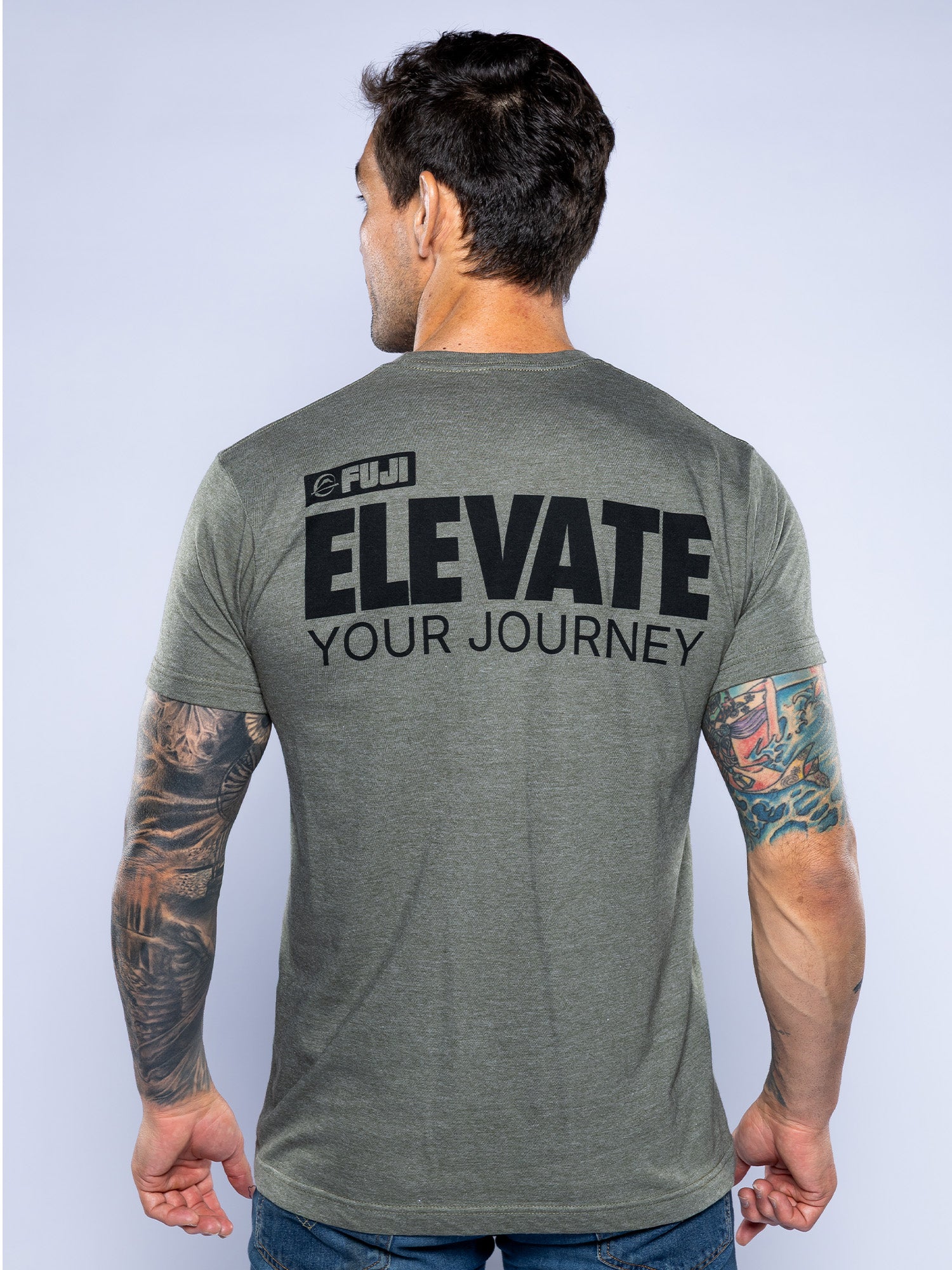 A man with short dark hair and tattoos on both arms is seen from behind wearing the Elevate Your Journey Shirt in heather grey, featuring "FUJI ELEVATE YOUR JOURNEY" printed in black on the back.