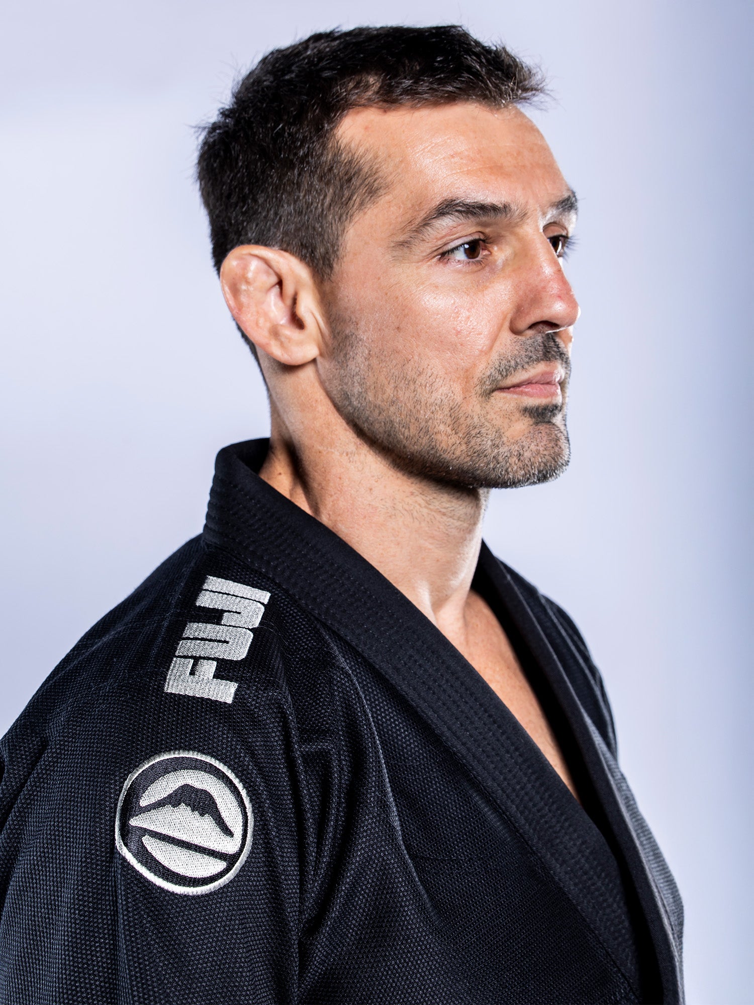 A man with short dark hair and light stubble wears the Baseline 2.0 BJJ Gi Black Grey, featuring white patches, as he looks to the side with a neutral expression against a plain, light background.