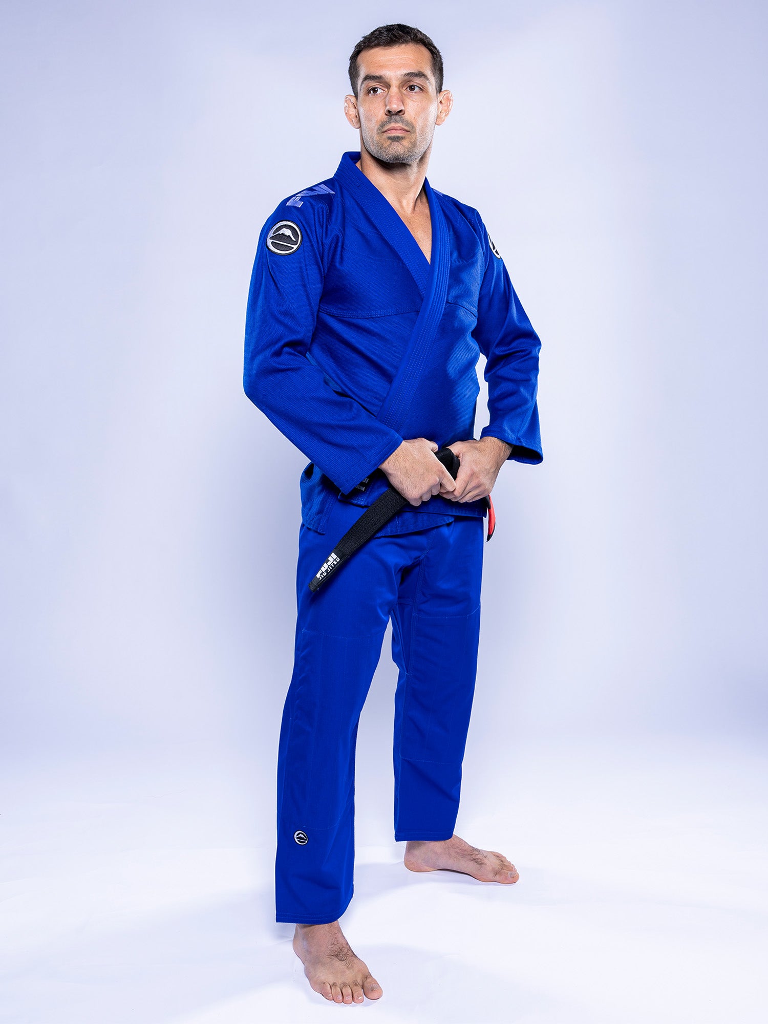 A man wears the Baseline 2.0 BJJ Gi Blue White, standing barefoot on a white background, confidently holding a black belt in front of him.