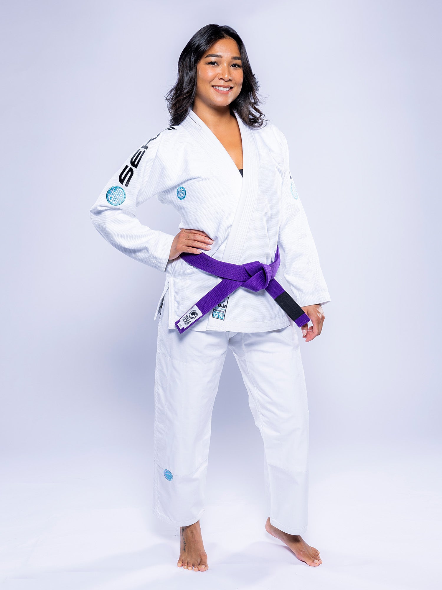 A woman with shoulder-length dark hair stands barefoot on a white background, smiling with her hands on her hips. She wears the Womens SEKAI 3.0 BJJ Gi White Teal and a purple belt tied at the waist.