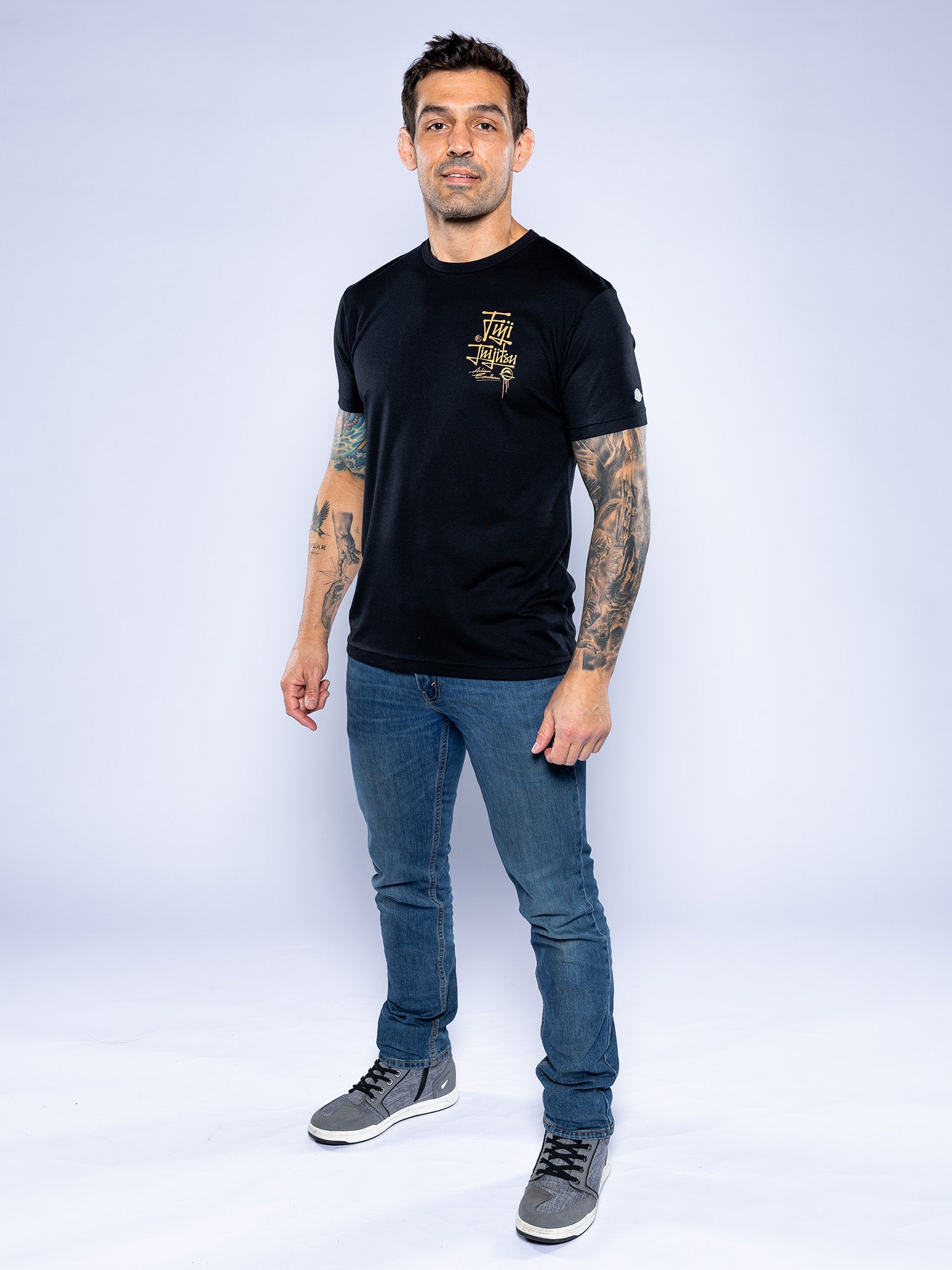 A man with tattooed arms stands and smiles slightly, wearing the Japanese Street Art Shirt Black, blue jeans, and gray sneakers against a plain light background.