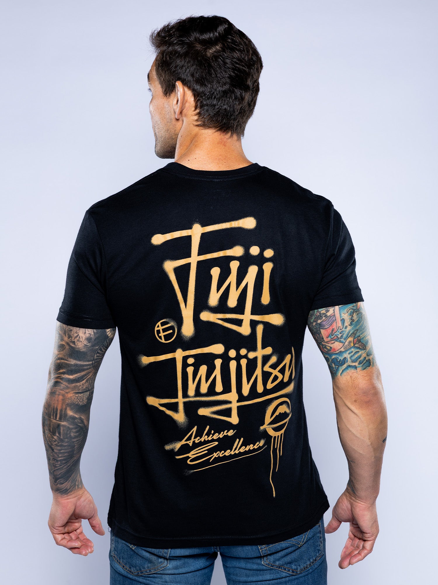A man with tattooed arms faces away, wearing the Japanese Street Art Shirt Black featuring gold graffiti-style "Fuji Jiu Jitsu Achieve Excellence" text on the back.