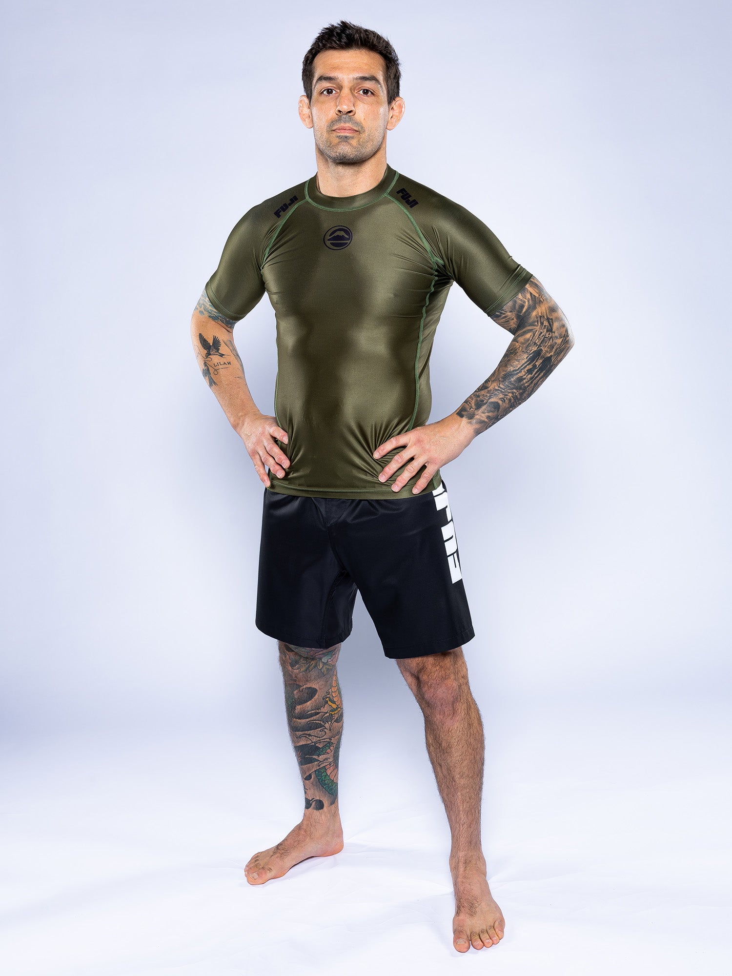 A tattooed man stands barefoot on a white background, confidently wearing the Baseline 2.0 Short Sleeve Rashguard in Military Green and black shorts with white lettering. He faces forward with hands on his hips.