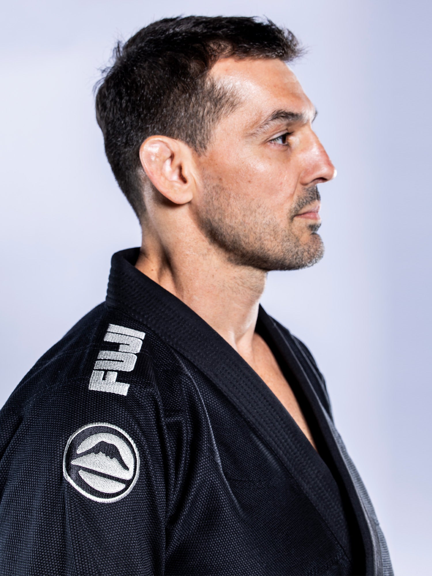 A man with short dark hair and a trimmed beard wears the Baseline 2.0 BJJ Gi Black Grey, featuring FUJI and a mountain logo on the shoulder, shown in profile against a plain light background.