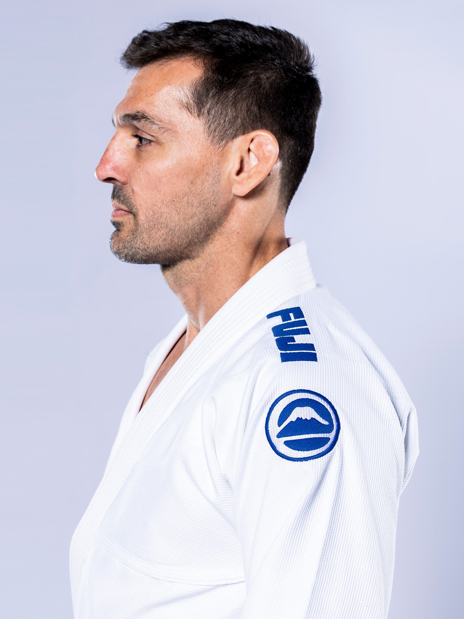 A man with short dark hair and stubble stands in profile, wearing the Baseline 2.0 BJJ Gi White Navy, which features blue patches and emblems on the shoulders. The background is light and plain.