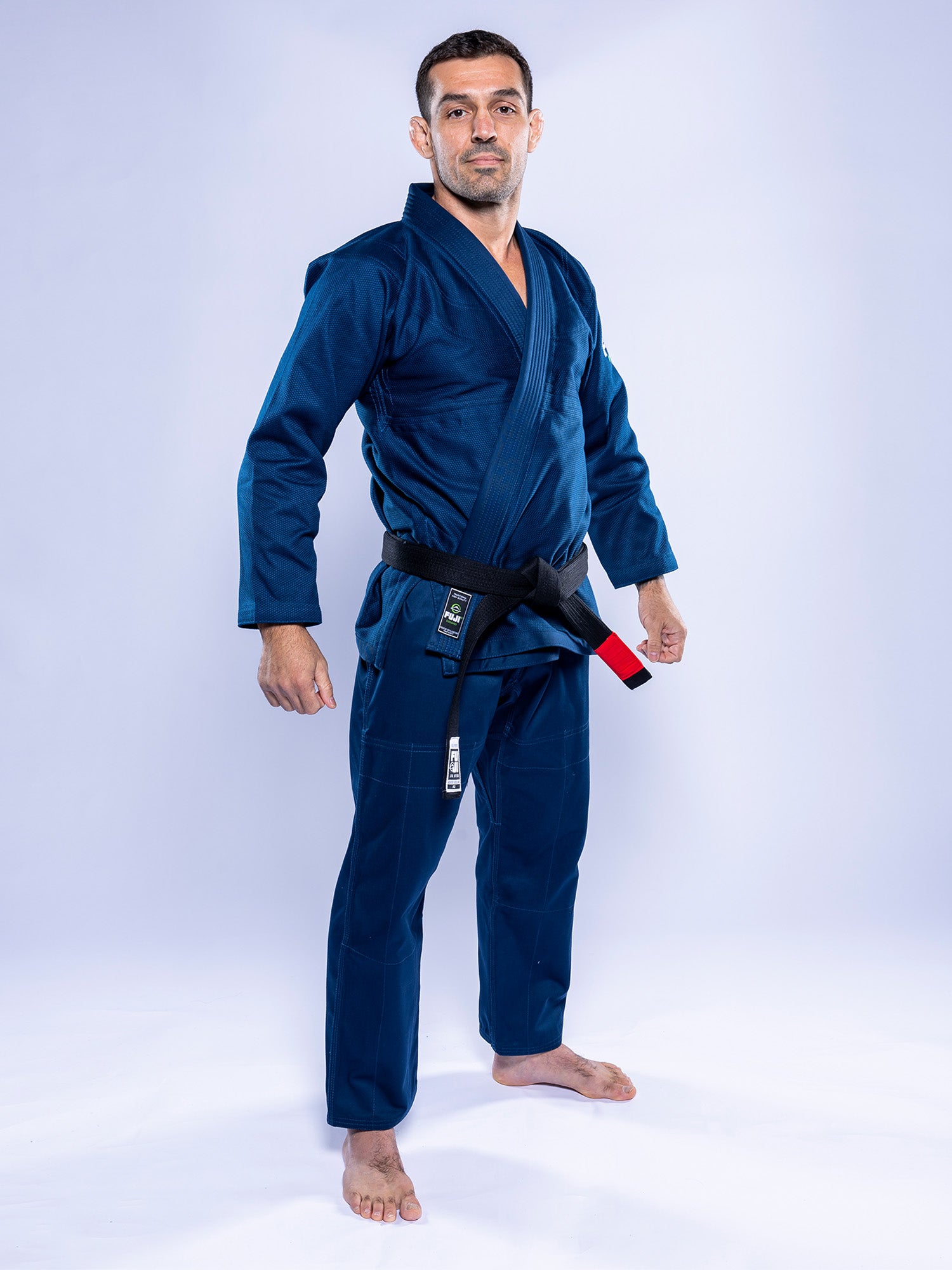A man stands barefoot on a white background wearing the Lightweight BJJ Gi Navy with a black belt. He faces forward, appearing confident and relaxed.