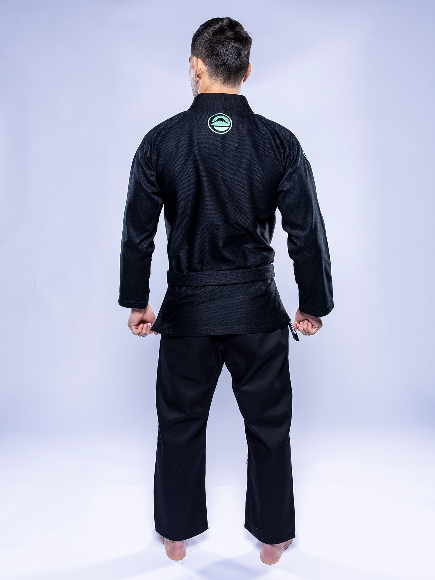 A person stands barefoot on a light surface, facing away from the camera, wearing the Baseline 2.0 BJJ Gi Black Mint, featuring a green circular patch on the back. The backdrop is plain and light-colored.