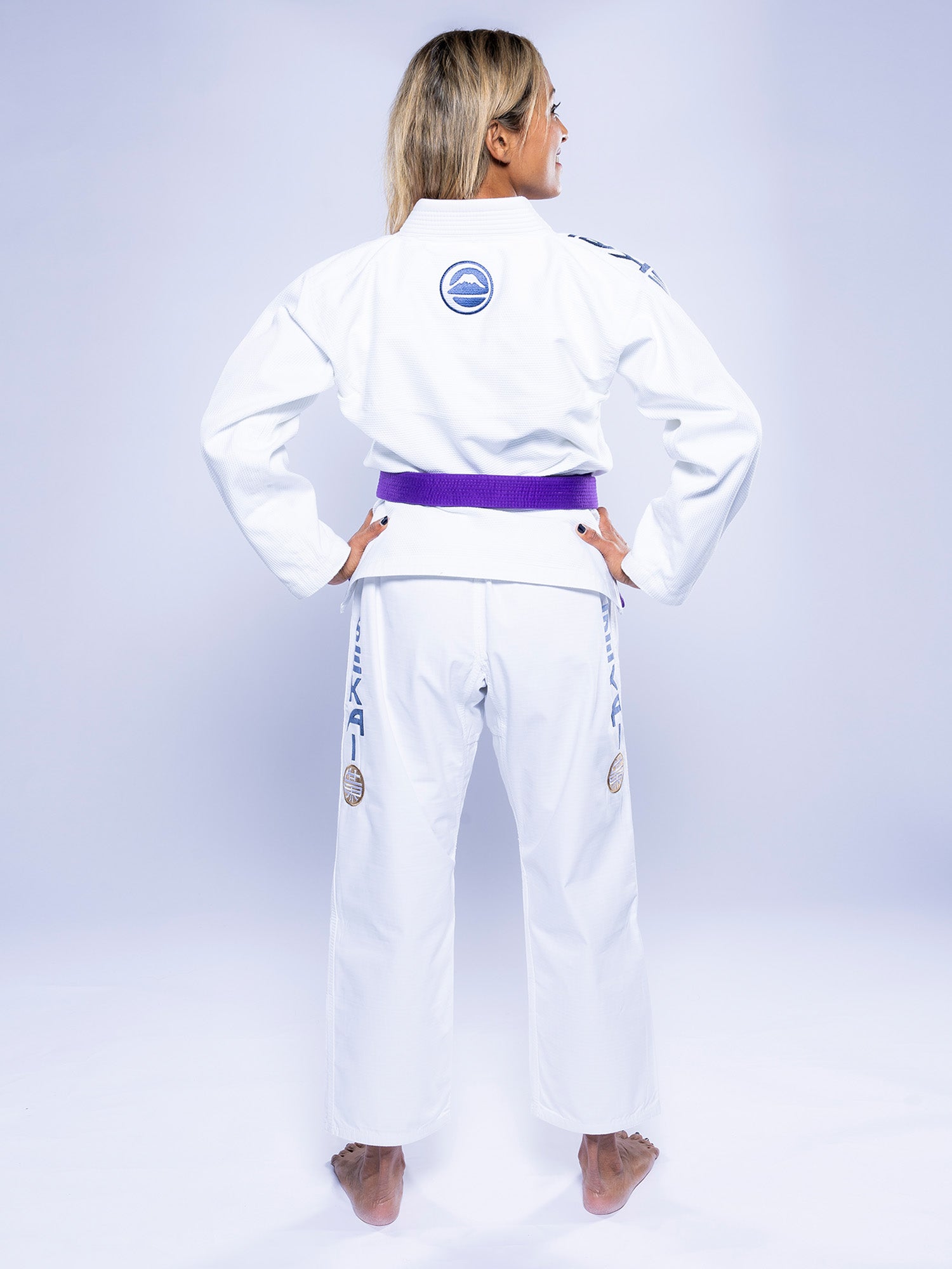 A person with long blonde hair faces away, wearing the Womens SEKAI 3.0 BJJ Gi White Navy with a purple belt. The gi features blue and gray patches and logos on the back and legs, set against a plain light background.