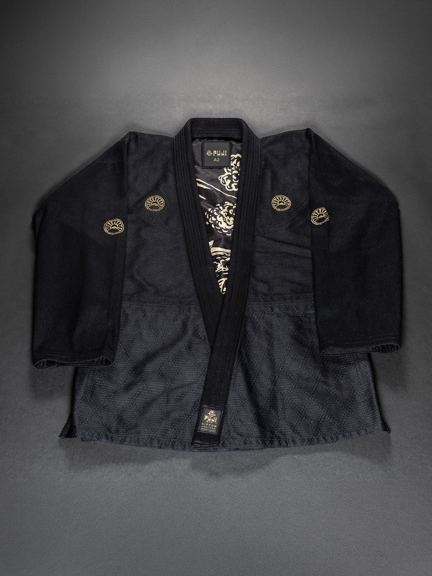 The Heritage Keisho Lightweight BJJ Gi Black is displayed flat on a dark background, featuring gold emblems and a patterned inner lining.