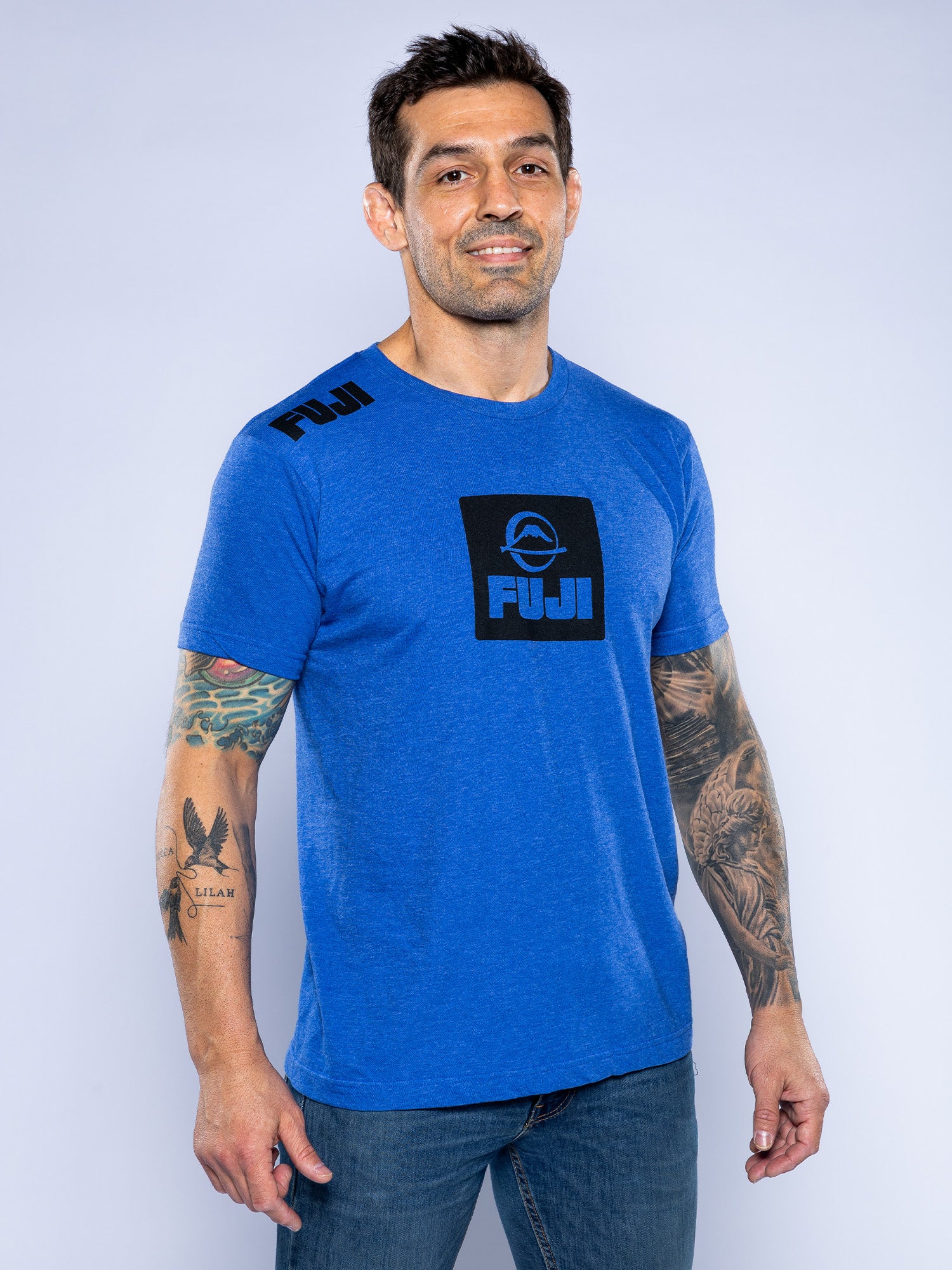 A man with short dark hair and tattoos on both arms smiles, wearing the Elevate Your Journey Shirt Blue and jeans. The background is plain and light-colored.