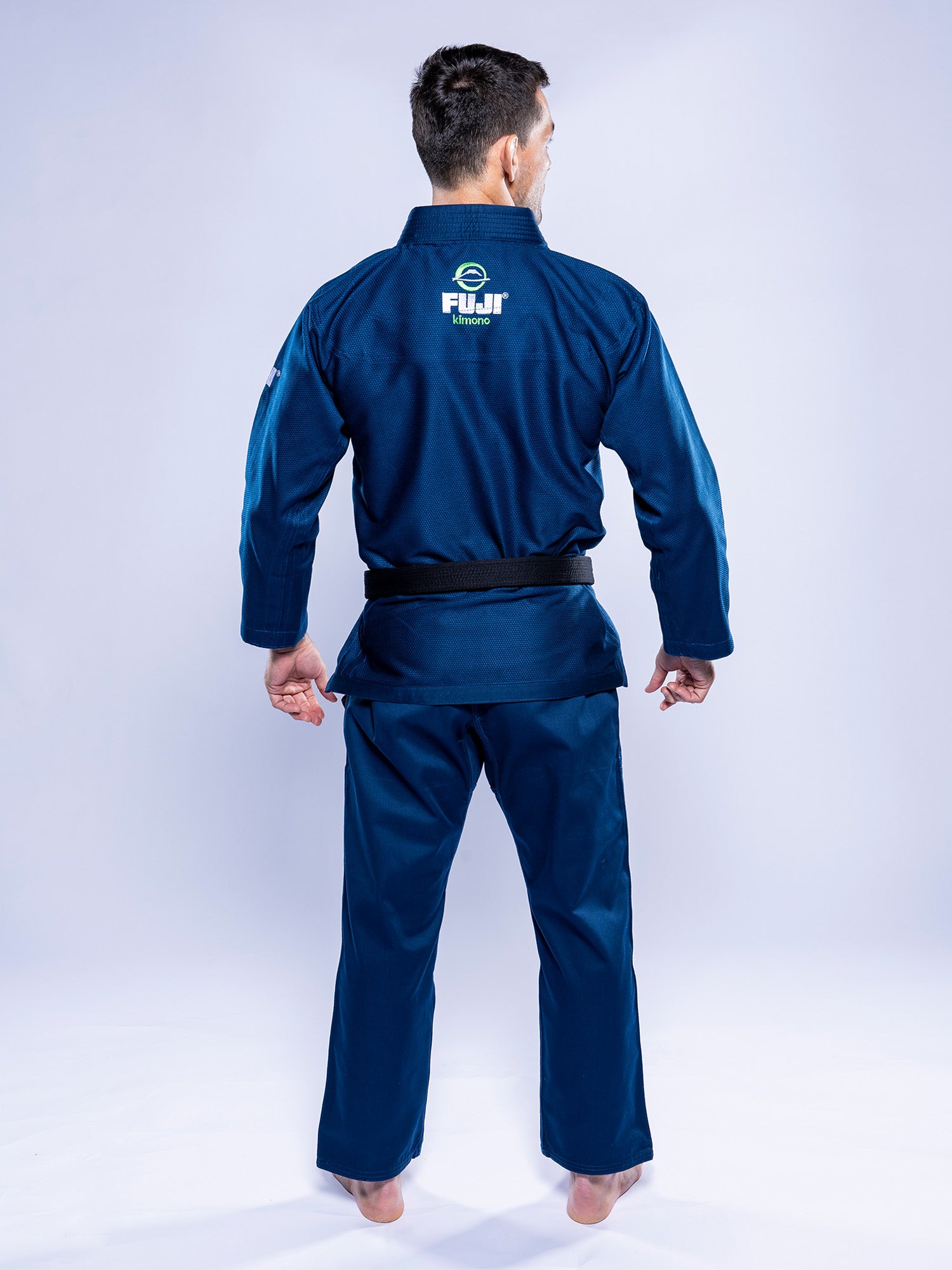 A man stands barefoot on a light background, facing away to reveal the FUJI logo in white on the back of his Lightweight BJJ Gi Navy.