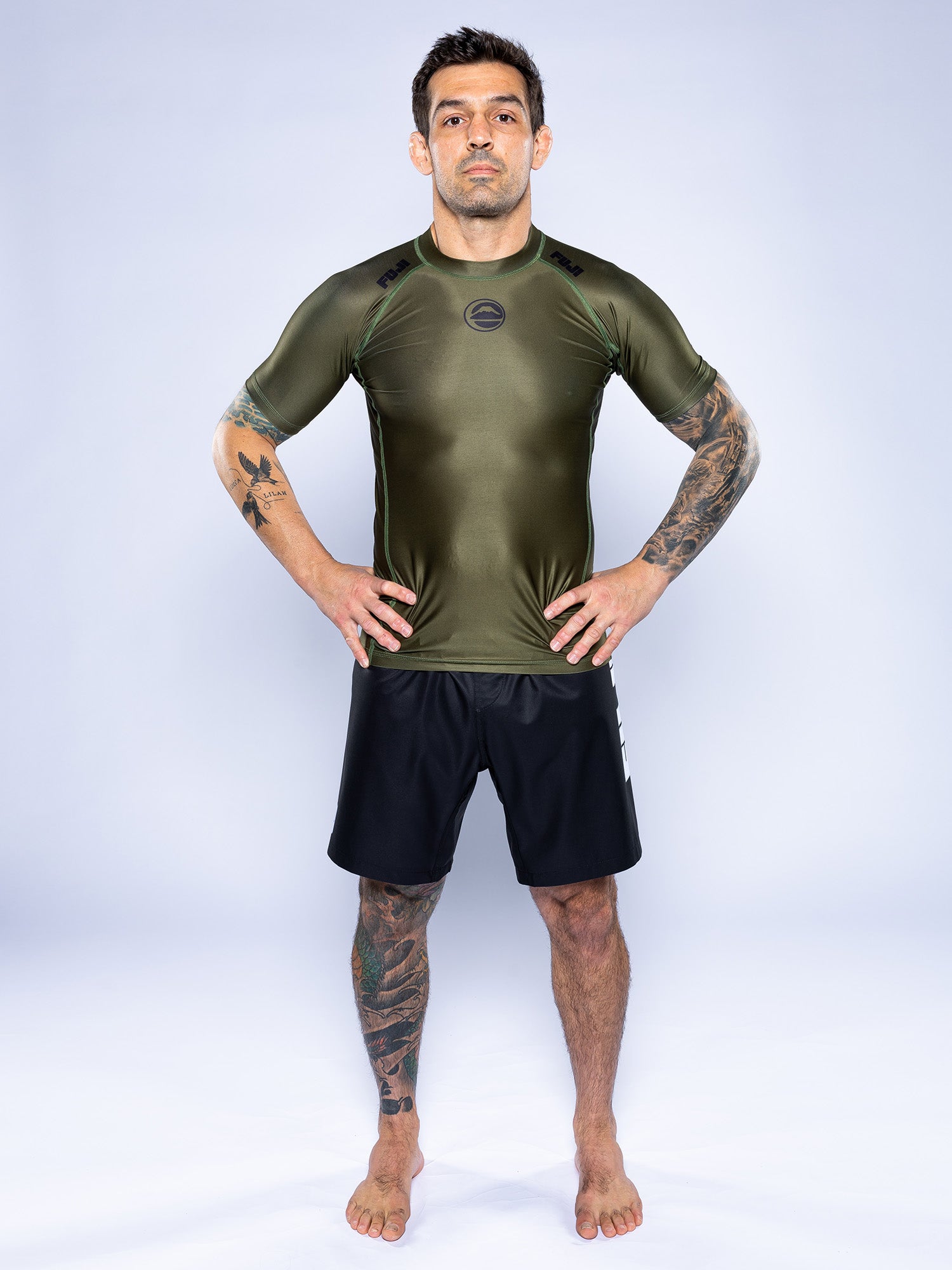 A man stands barefoot on a light background, wearing the Baseline 2.0 Short Sleeve Rashguard in Military Green and black athletic shorts. He has tattoos on both arms and one leg, with his hands resting on his hips.