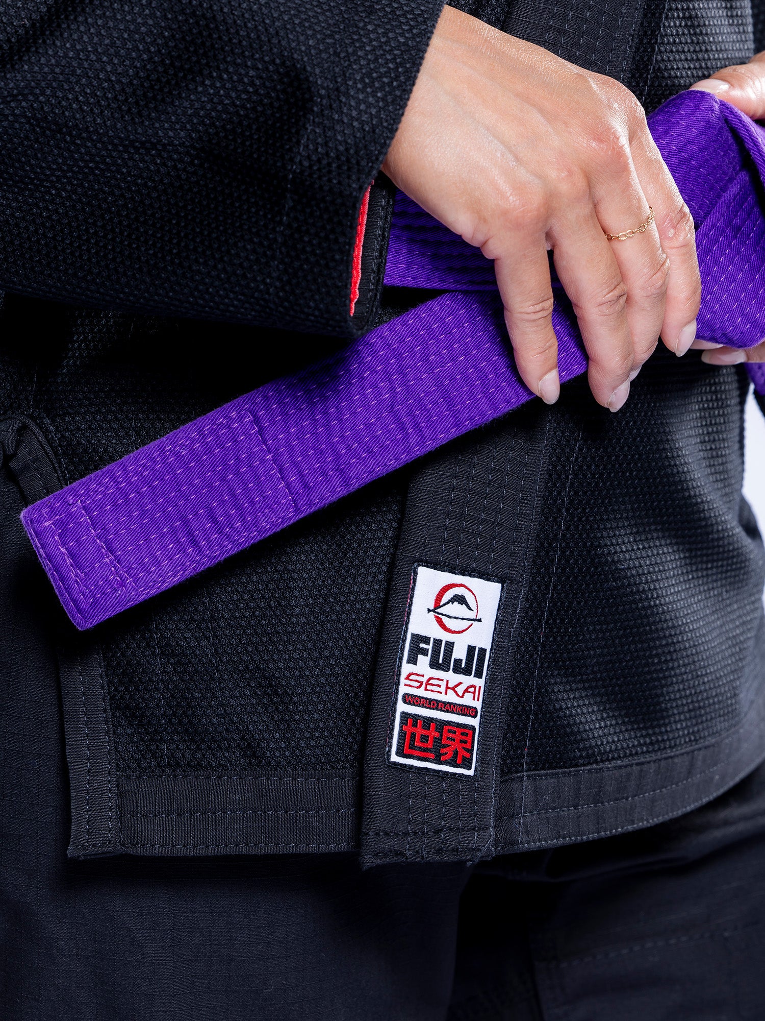 Close-up of a person’s hands tying a purple belt around the waist of the Womens SEKAI 3.0 BJJ Gi Black Grey, featuring a Fuji Seki patch with Japanese characters.