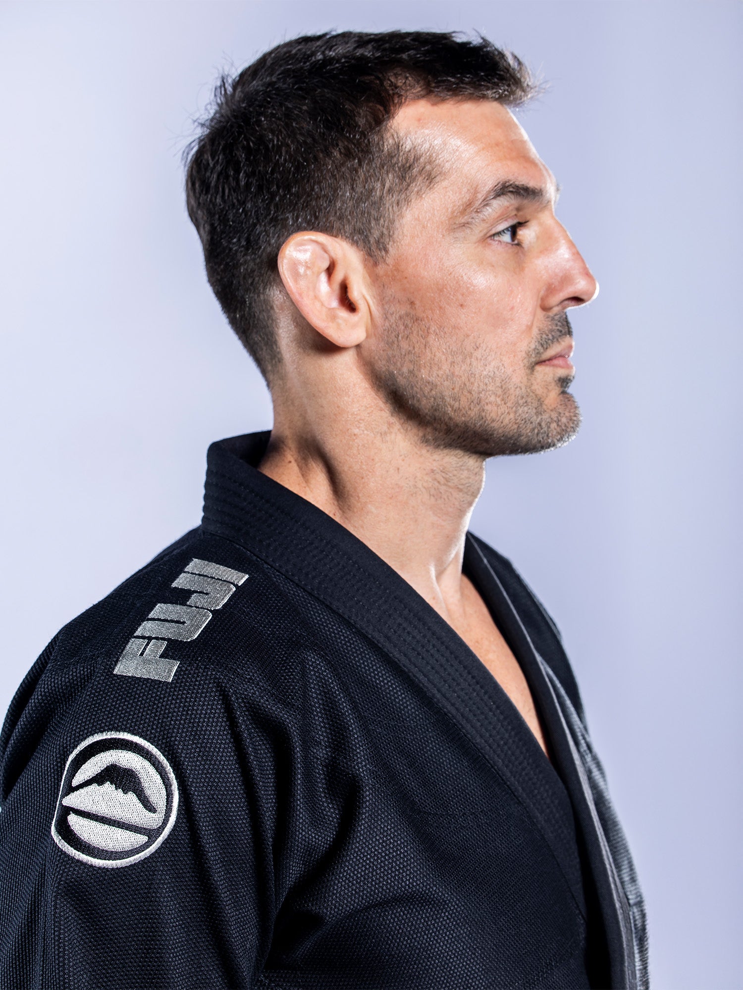 A man with short dark hair stands in profile against a light background, wearing the Baseline 2.0 BJJ Gi Black Grey, which features FUJI logos on the shoulder and chest.