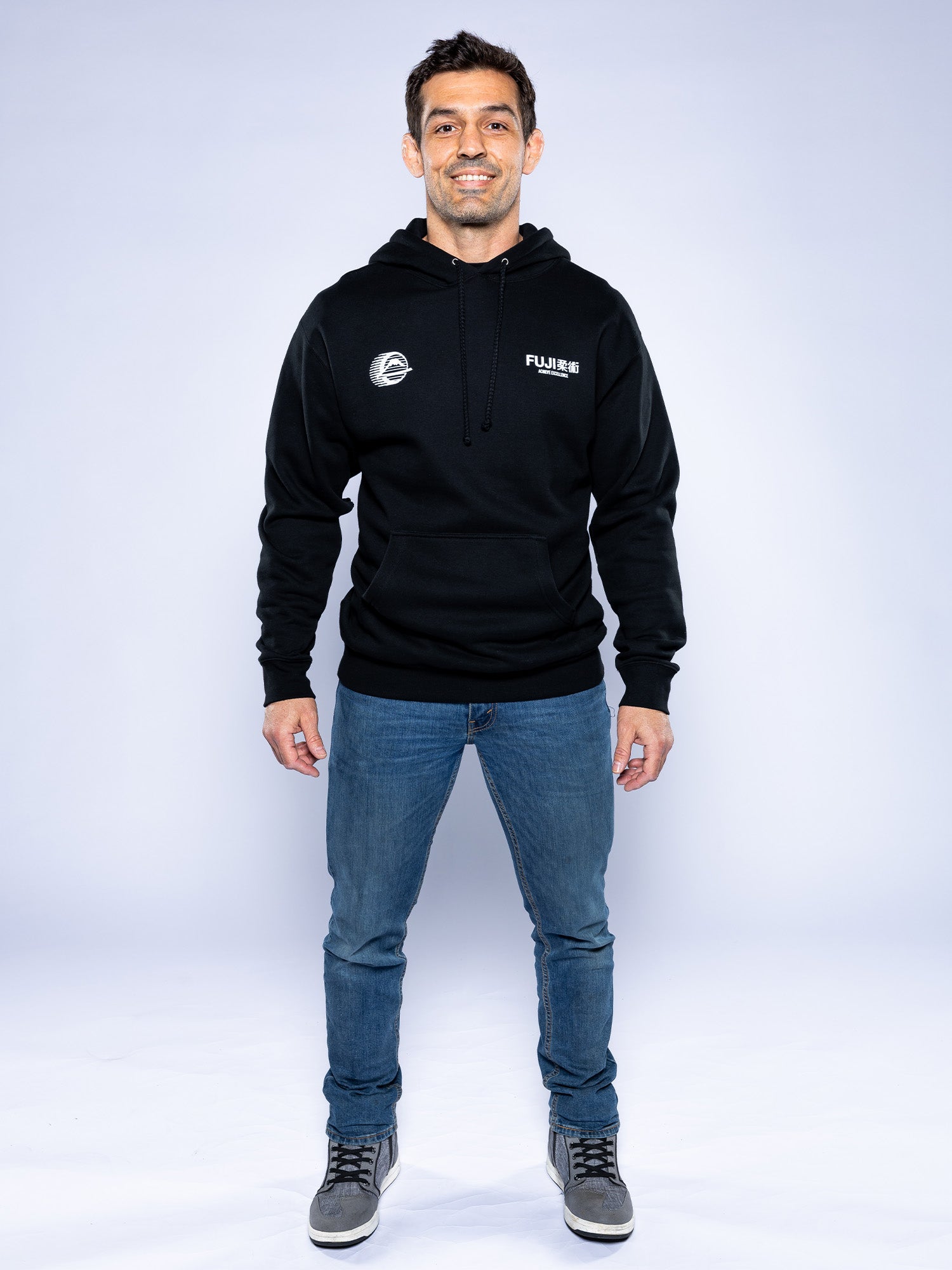 A man stands facing forward, smiling, wearing the Iconic Jiu Jitsu Hoodie Black with white chest logos, blue jeans, and gray sneakers against a plain light background.