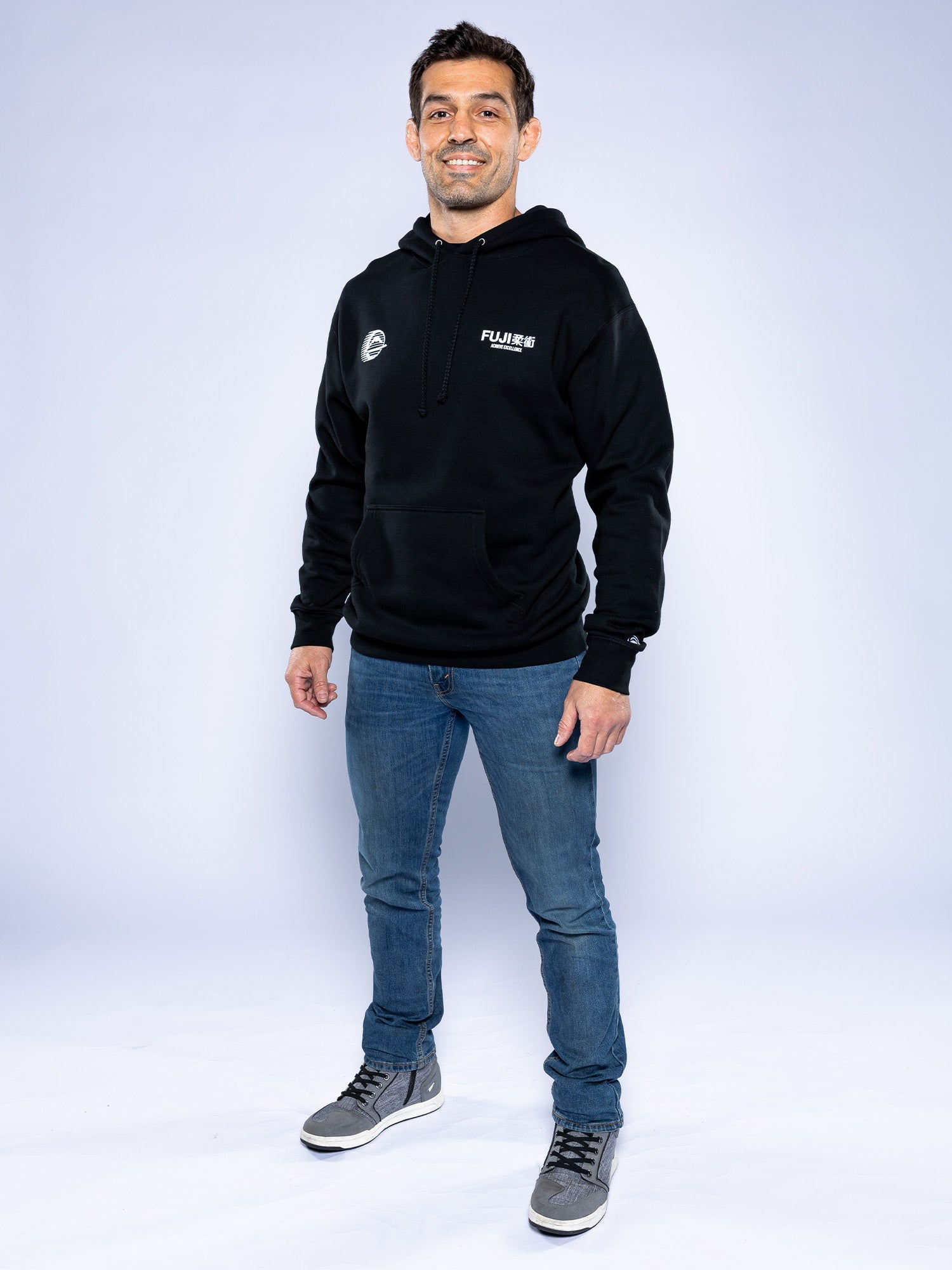 A man stands smiling in the Iconic Jiu Jitsu Hoodie Black, paired with blue jeans and gray high-top sneakers against a plain light background.