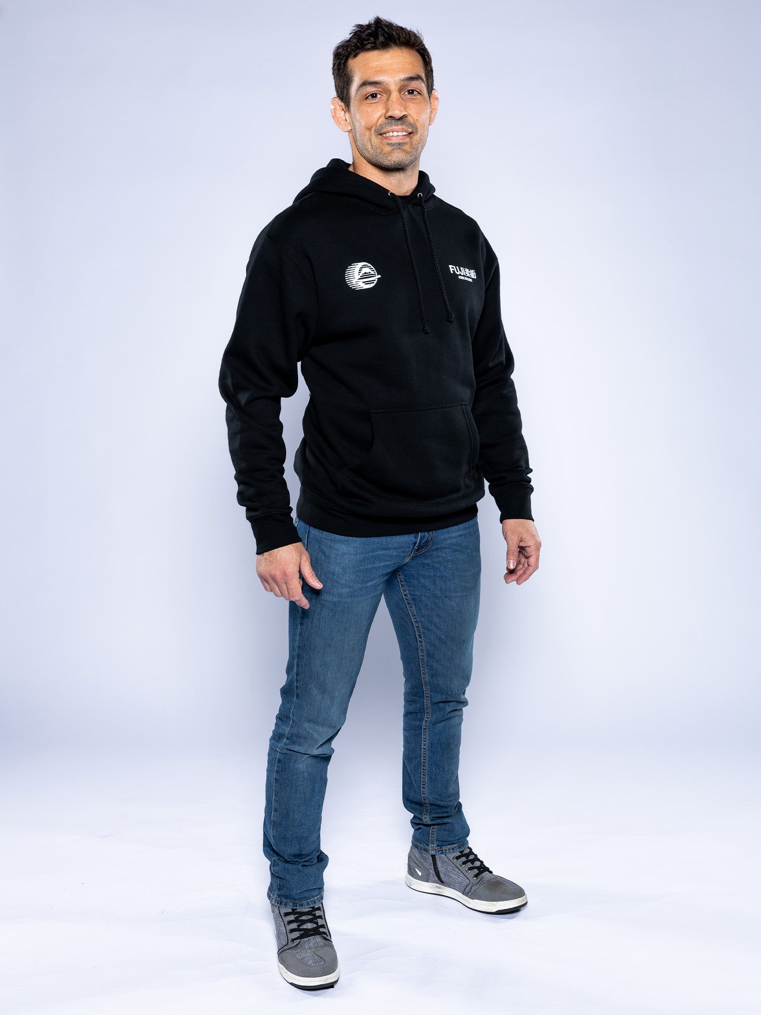 A man smiles against a plain light background, wearing the Iconic Jiu Jitsu Hoodie Black, blue jeans, and gray sneakers.