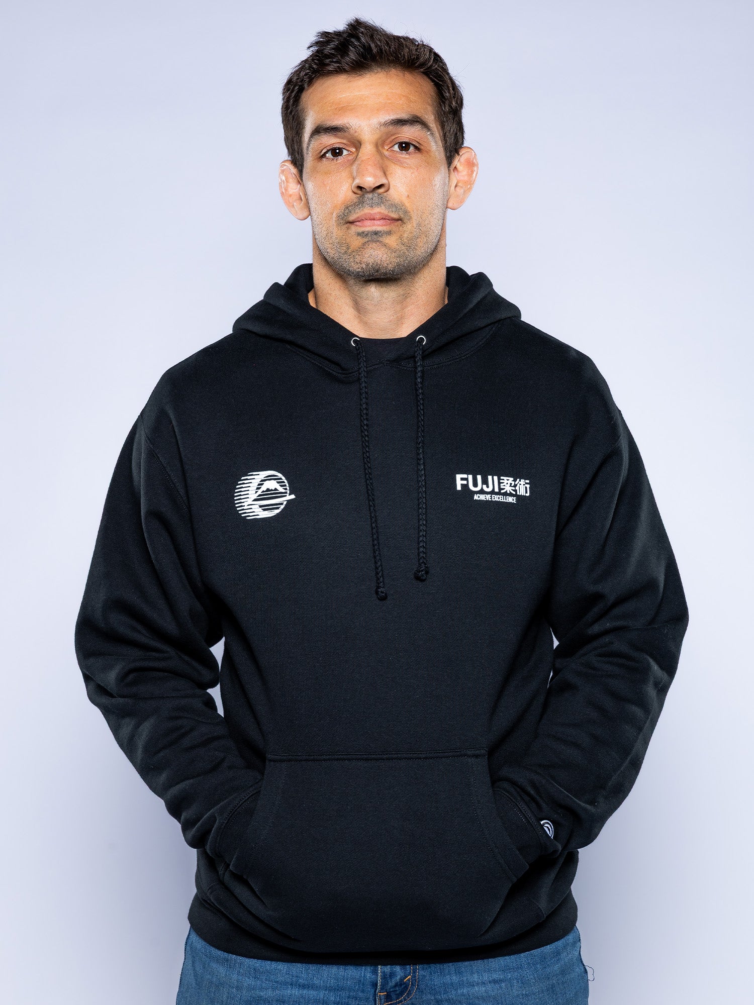 A man with short dark hair and a neutral expression stands facing forward, hands in the pocket of the Iconic Jiu Jitsu Hoodie Black, featuring white logos, against a plain light background.