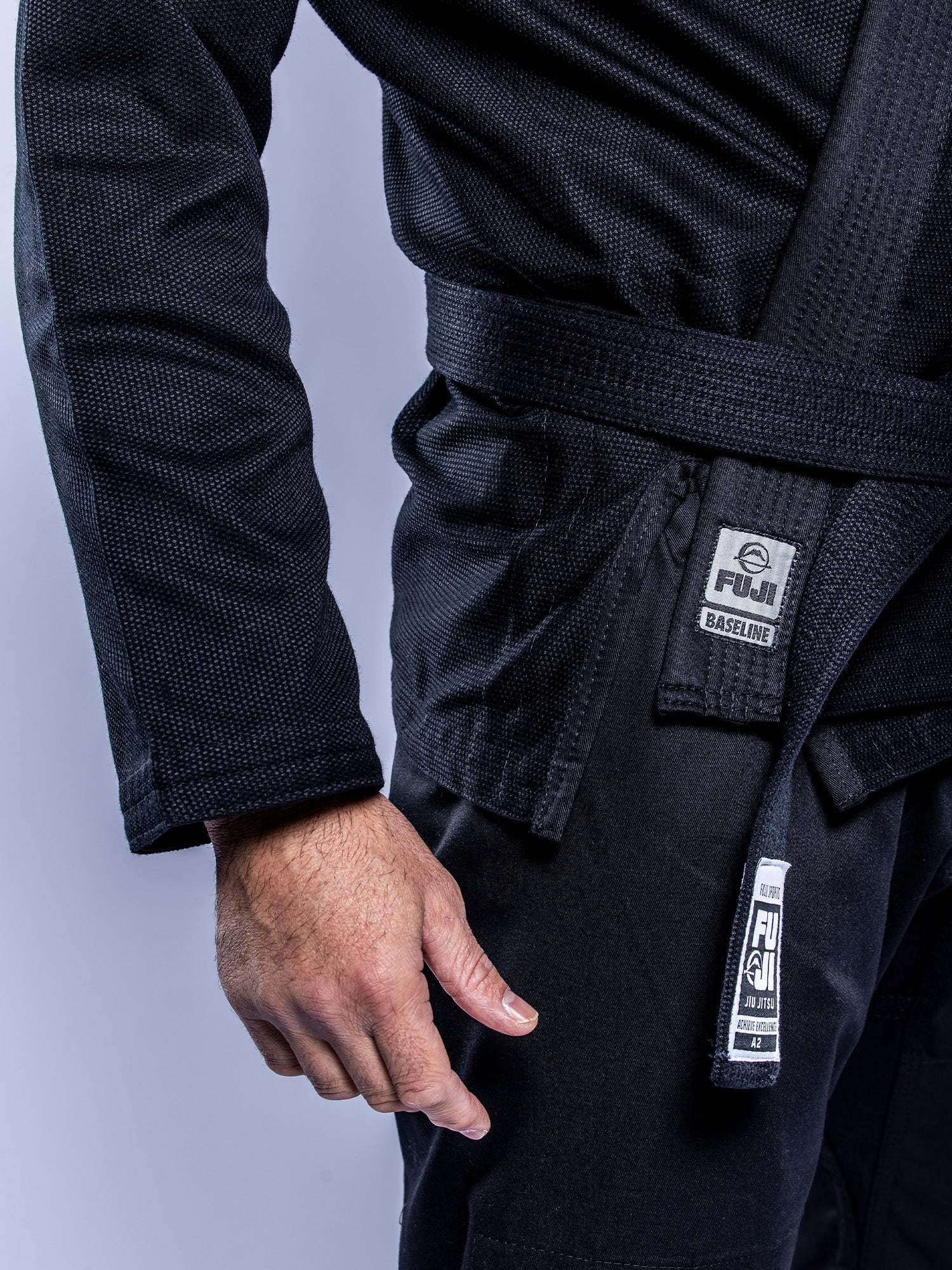 A person is shown from the chest down, one arm relaxed by their side, wearing a black Baseline 2.0 BJJ Gi Black Grey and a black belt against a plain background.