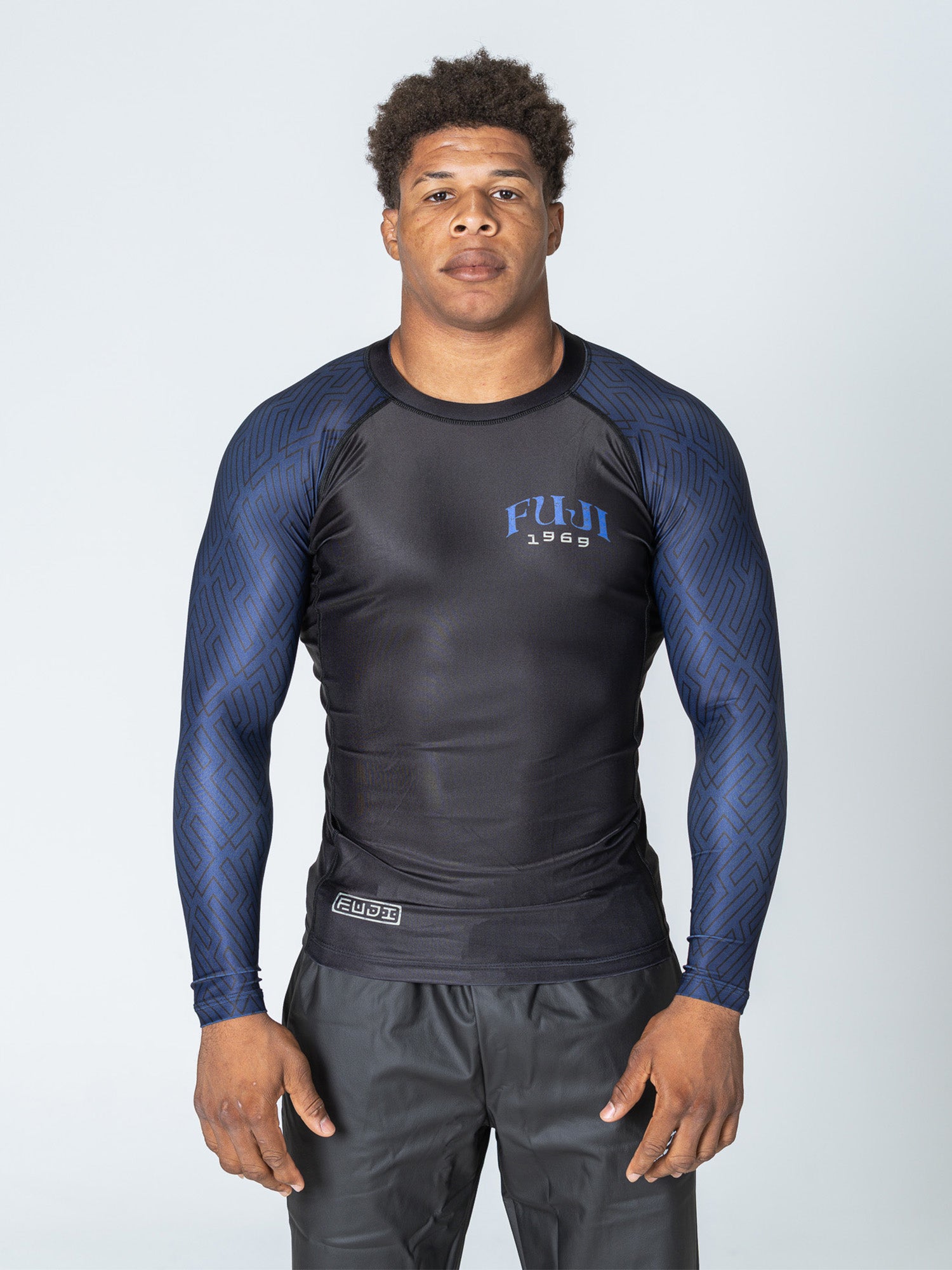 A man wearing the Oni Samurai Flex Lite Rashguard Blue and black pants stands facing forward with a neutral expression against a plain light background.
