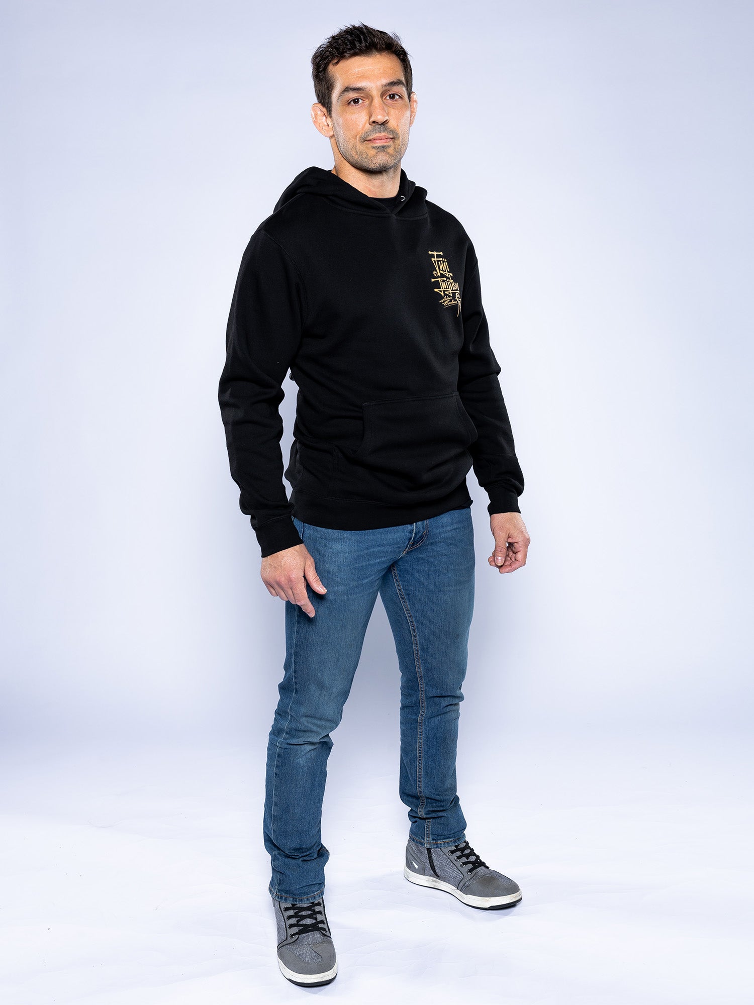 A man stands against a light background wearing the Japanese Street Art Hoodie Black, paired with blue jeans and gray high-top sneakers. He faces slightly to the side with a neutral expression and relaxed hands at his sides.