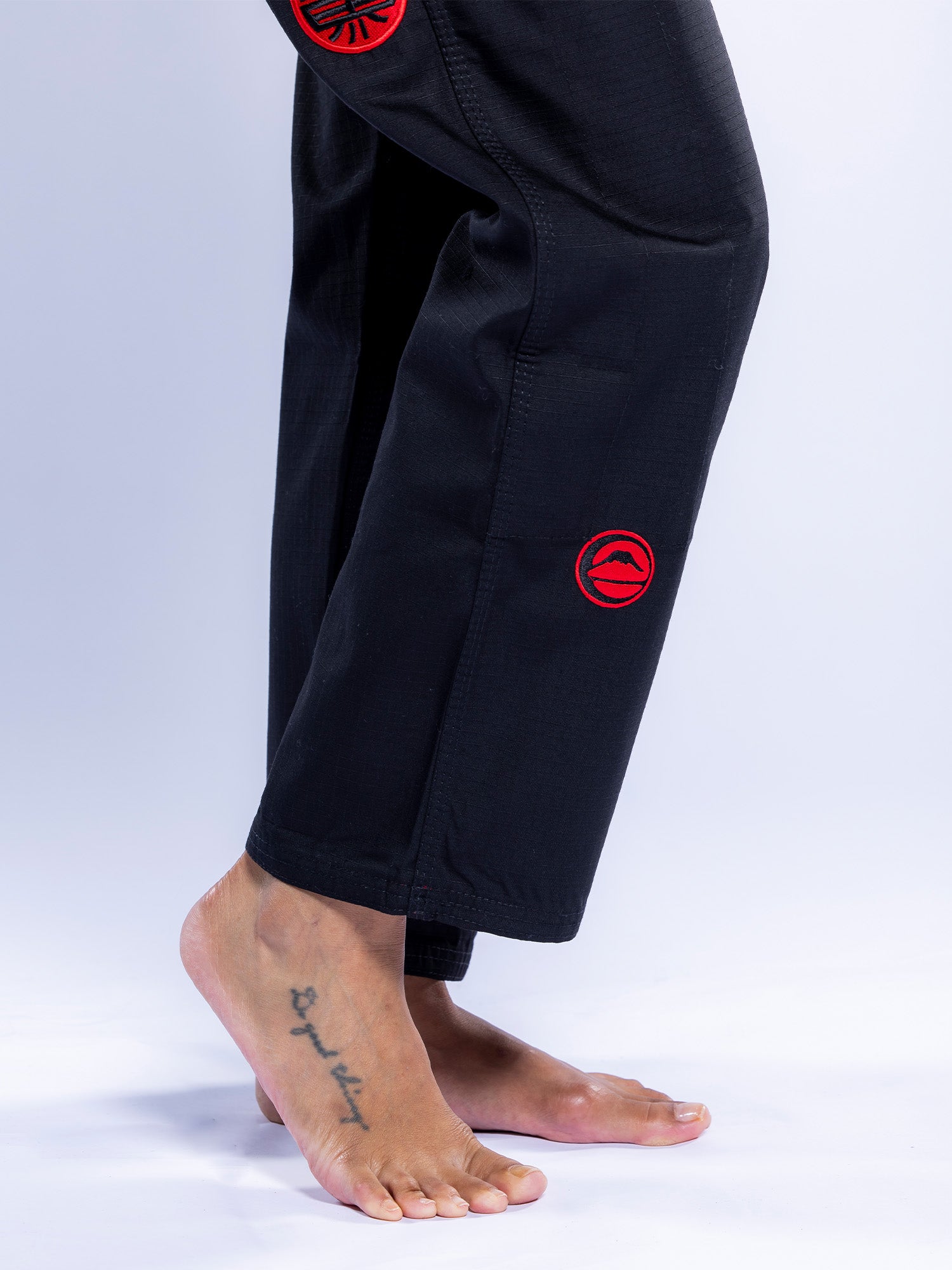 A person stands barefoot on tiptoes in the Womens SEKAI 3.0 BJJ Gi Black Grey, featuring red circular embroidered patches. A star tattoo is visible on the outer left foot against a plain white background.