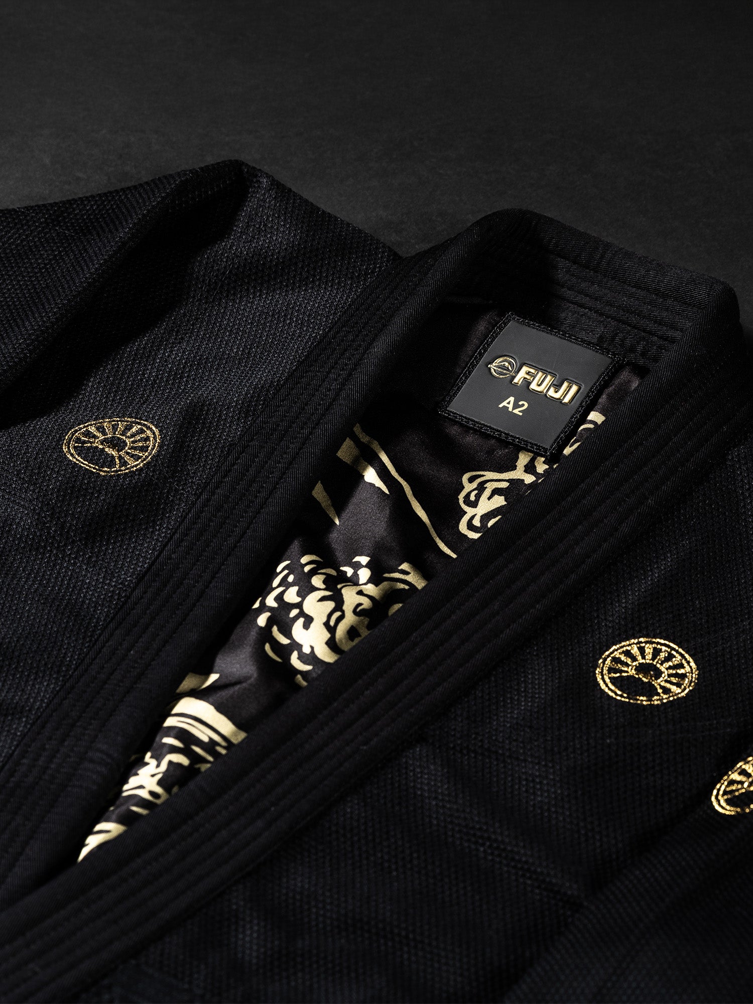 Close-up of the Womens Heritage Keisho Lightweight BJJ Gi in black, featuring gold embroidered patches and a patterned inner lining, displayed on a dark surface.