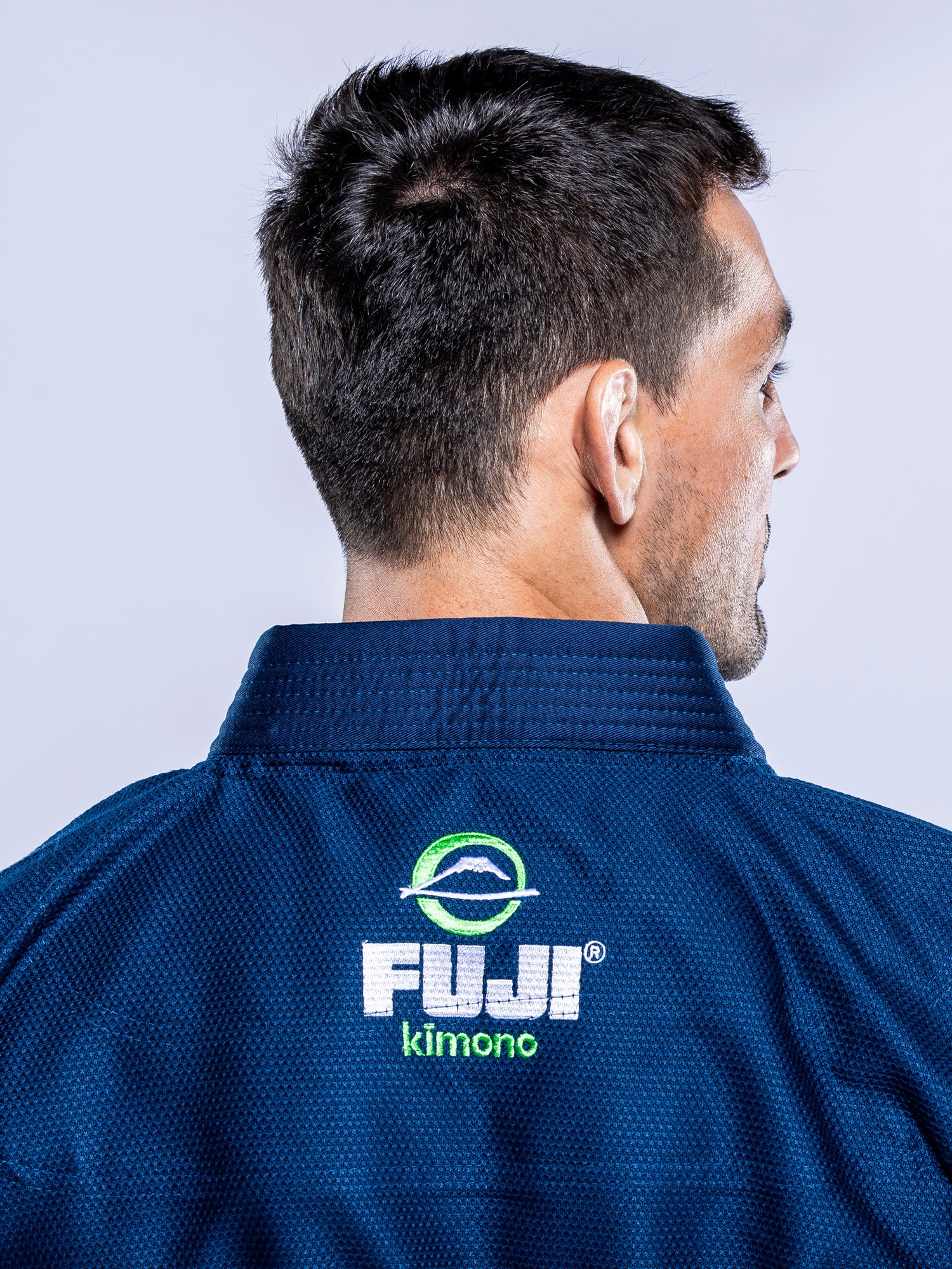 A man is seen from behind wearing a Lightweight BJJ Gi Navy with the brand logo and name displayed on the back collar. The background is light and neutral.