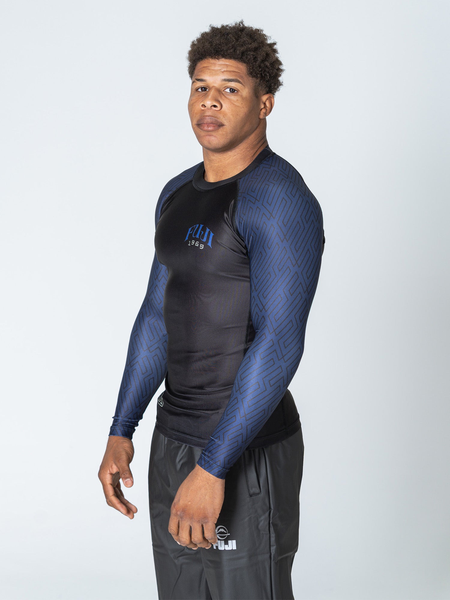 A man with short curly hair wears the Oni Samurai Flex Lite Rashguard Blue and black sports pants, standing slightly to the side against a plain light background.
