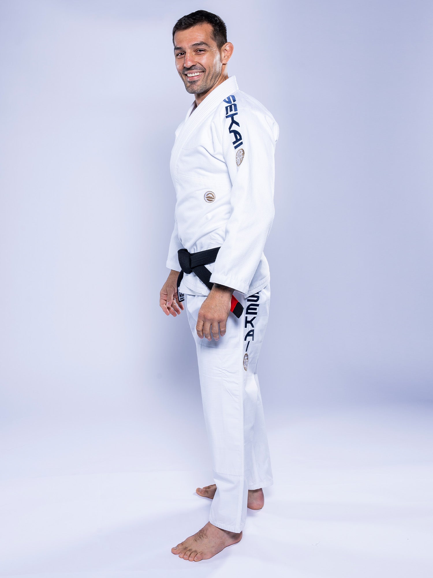 A man wearing the SEKAI 3.0 BJJ Gi White Navy with dark lettering on the sleeve and pants stands barefoot on a white background, smiling and looking at the camera.