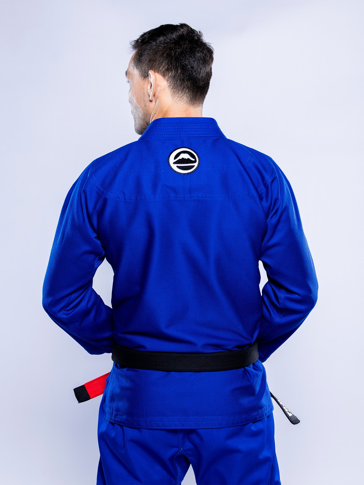 A man stands with his back to the camera, hands behind him, wearing the Baseline 2.0 BJJ Gi Blue White with a white and black circular patch on the upper back. The background is plain light gray.