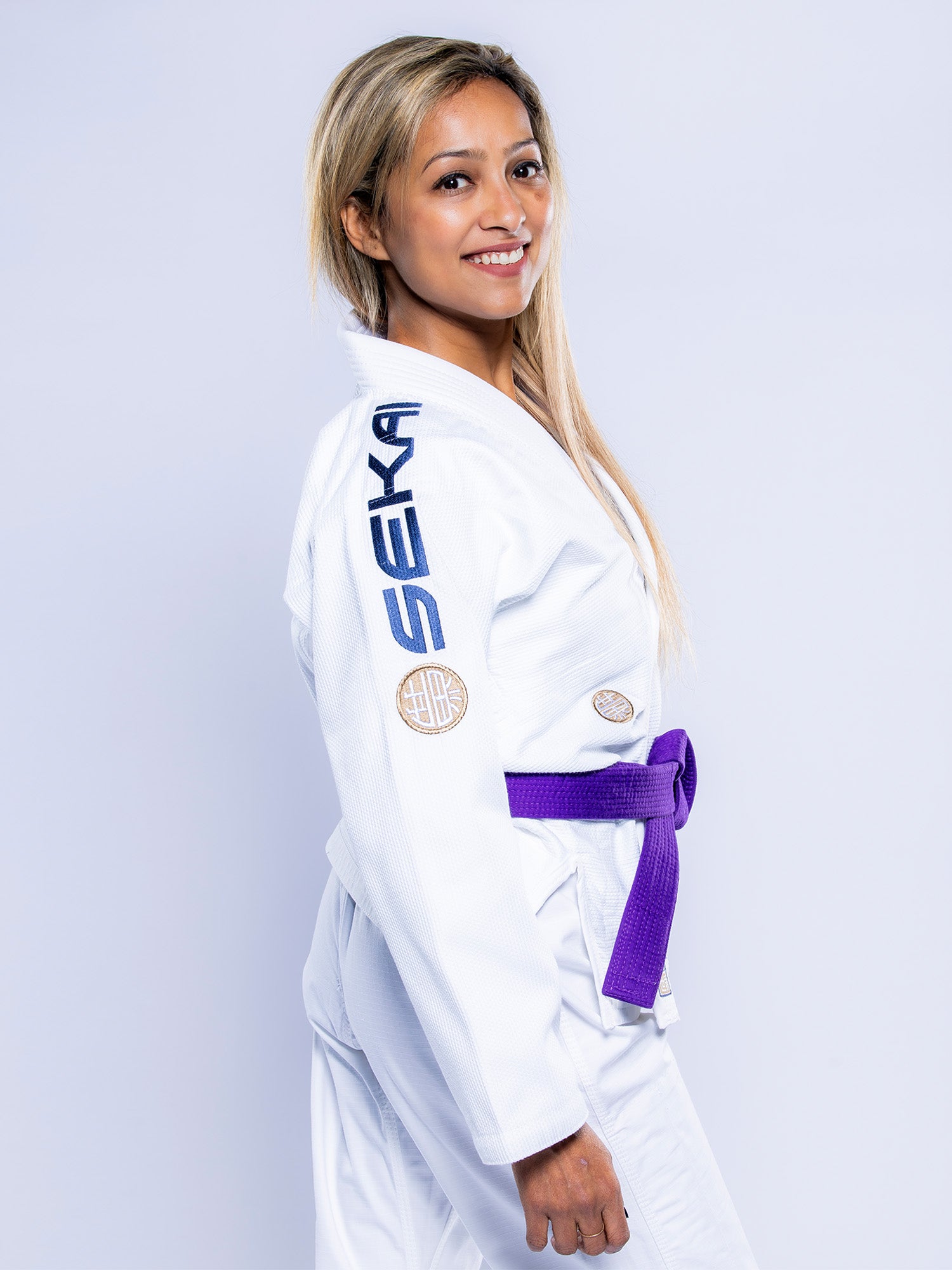 A woman with long blonde hair smiles while wearing the Womens SEKAI 3.0 BJJ Gi White Navy with a purple belt. SEKA is written on the shoulder, and patches are on the chest and arm. The background is plain light gray.