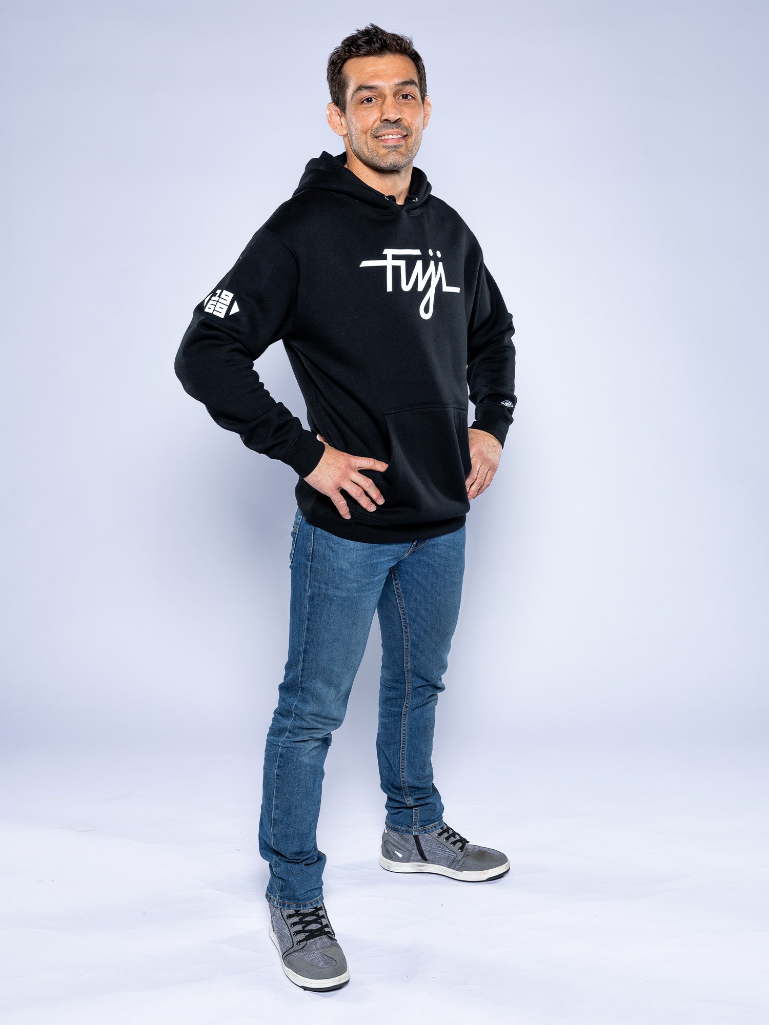 A man stands confidently with hands on hips, wearing the Midnight Collegiate Pull Over Hoodie Black, blue jeans, and gray sneakers against a light gray studio background.