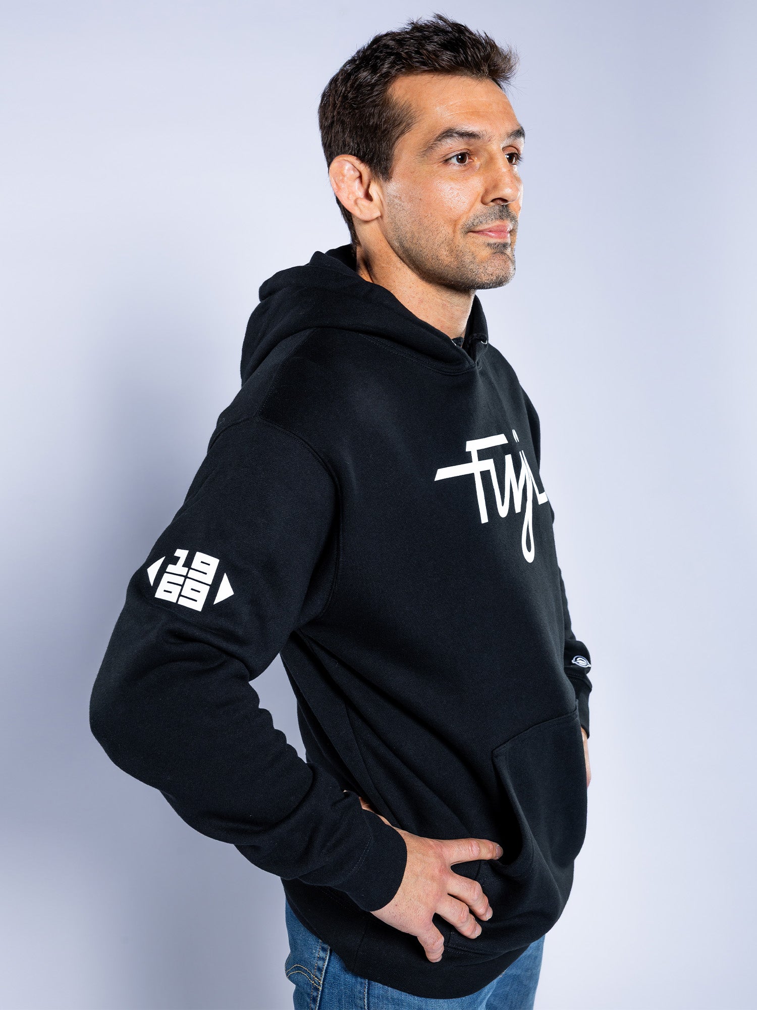 A man stands in profile against a light background, wearing the Midnight Collegiate Pull Over Hoodie Black with white lettering on the chest and "1969" on the sleeve, hands on hips and looking slightly to the side.