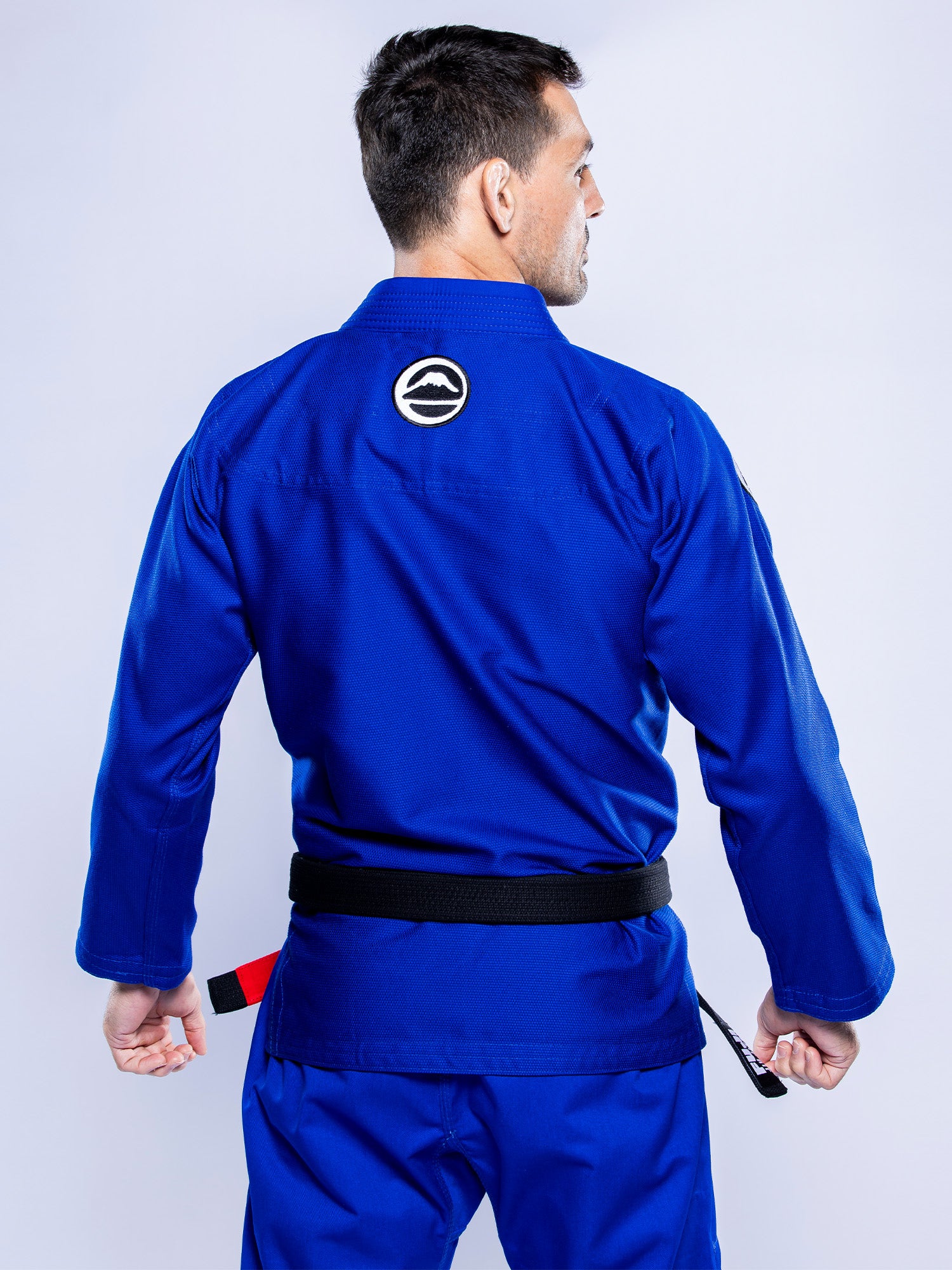 A man in the Baseline 2.0 BJJ Gi Blue White with a black belt stands with his back to the camera, holding his belt ends. The gi features a circular black and white patch on the upper back.