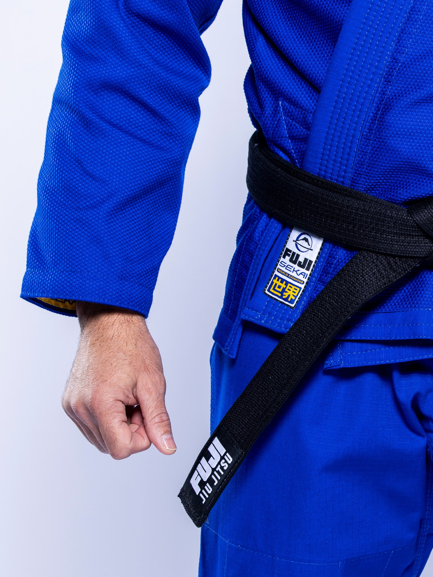 A person wearing the SEKAI 3.0 BJJ Gi Blue Yellow with a black belt labeled “FUJI JIU JITSU,” showing their fist and part of their torso.