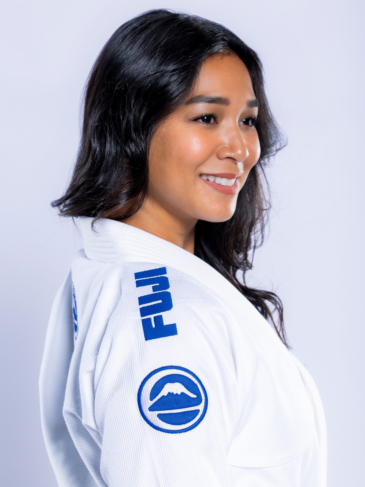 A woman with long dark hair smiles while wearing the Womens Baseline 2.0 BJJ Gi in White Navy, featuring blue FUJI logos on the collar and sleeve, standing against a light background.